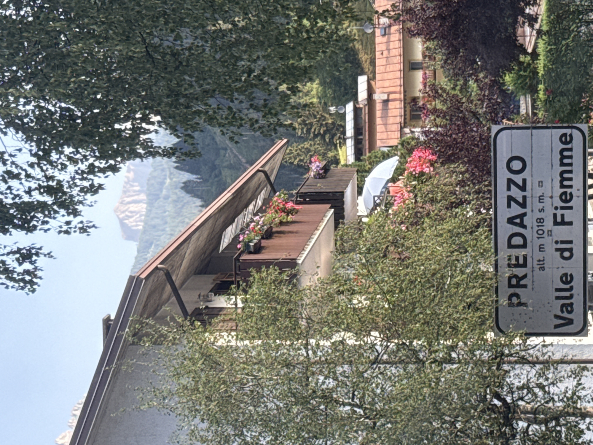 The image shows a street sign in Predazzo, Valle di Fiemme, Italy, in the Dolomite mountains. The sign reads "PREDAZZO" in large letters, with "alt. m 1018 s.m." and "Valle di Fiemme" below. The sign is white with black lettering and has a gray border. Behind the sign, a building with a balcony adorned with flowers is visible. The surrounding environment is characterized by lush greenery, trees, and distant mountains under a clear sky. The scene appears to be a typical mountain village setting.