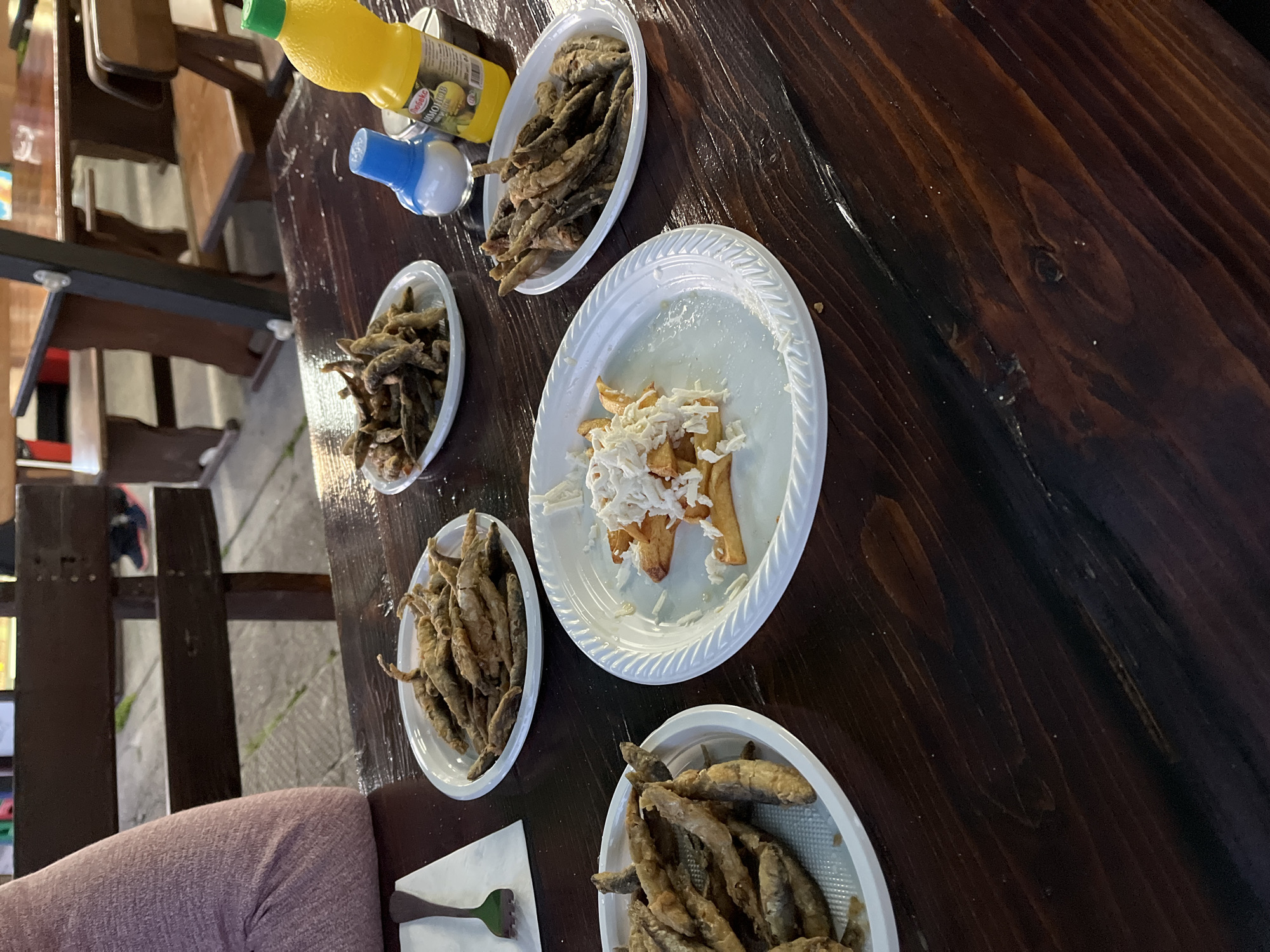 A close-up, eye-level, medium shot shows several white plastic plates filled with fried fish and french fries on a dark wooden table. The table is outdoors, with a blurred background of wooden benches and tables. The plates are arranged in a semi-circle, with a bottle of yellow liquid, a salt shaker with a blue lid, and a blue-lidded container of what appears to be tartar sauce or a similar condiment situated between them. One plate in the foreground contains french fries topped with shredded white cheese. A person's arm, clad in a pink knitted garment, is visible on the left side of the image. The setting appears to be a casual dining establishment in Smolyan, Bulgaria, given the presence of traditional Bulgarian cuisine and the overall ambiance.