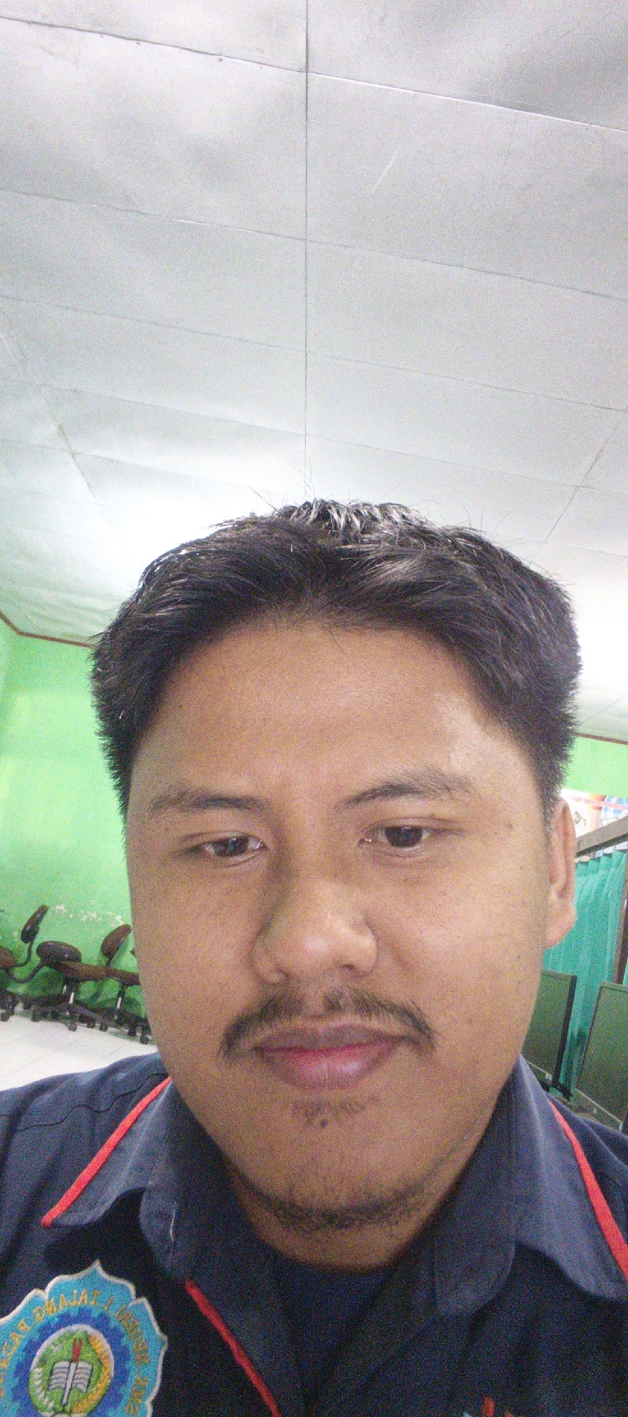 A medium shot selfie features a man with dark hair and a mustache. He is wearing a dark blue collared shirt with red trim and an emblem on the left chest. The man is looking directly at the camera with his eyes open. Behind him, a bright green wall is visible, and further back, several office chairs on wheels are arranged in a row. The ceiling appears to be made of white acoustic tiles. The image quality is grainy, suggesting it was taken in low light conditions. The emblem on the shirt displays text and imagery that appears to be a seal or logo, with a book and a torch prominently featured. The overall setting suggests an indoor environment, likely an office or classroom, possibly in Sukabumi, Indonesia, given the presence of what might be an Indonesian educational or institutional logo.