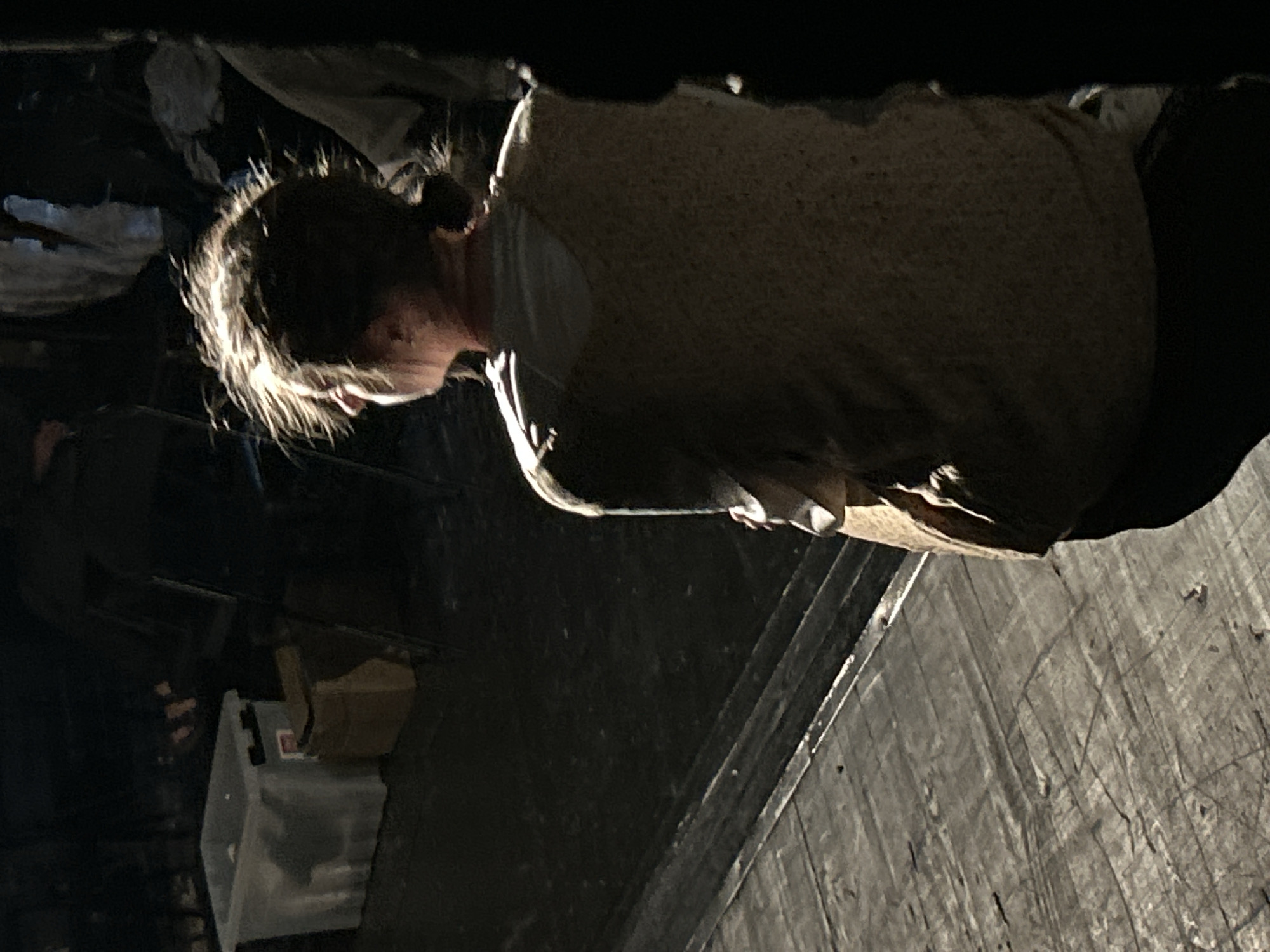 A person is seen from a low angle, head tilted downward. They wear a textured top and dark pants. The surroundings include a dark, cluttered area with storage containers and possibly stage equipment, and a planked surface, possibly a stage floor. The location is not discernible from the provided information.