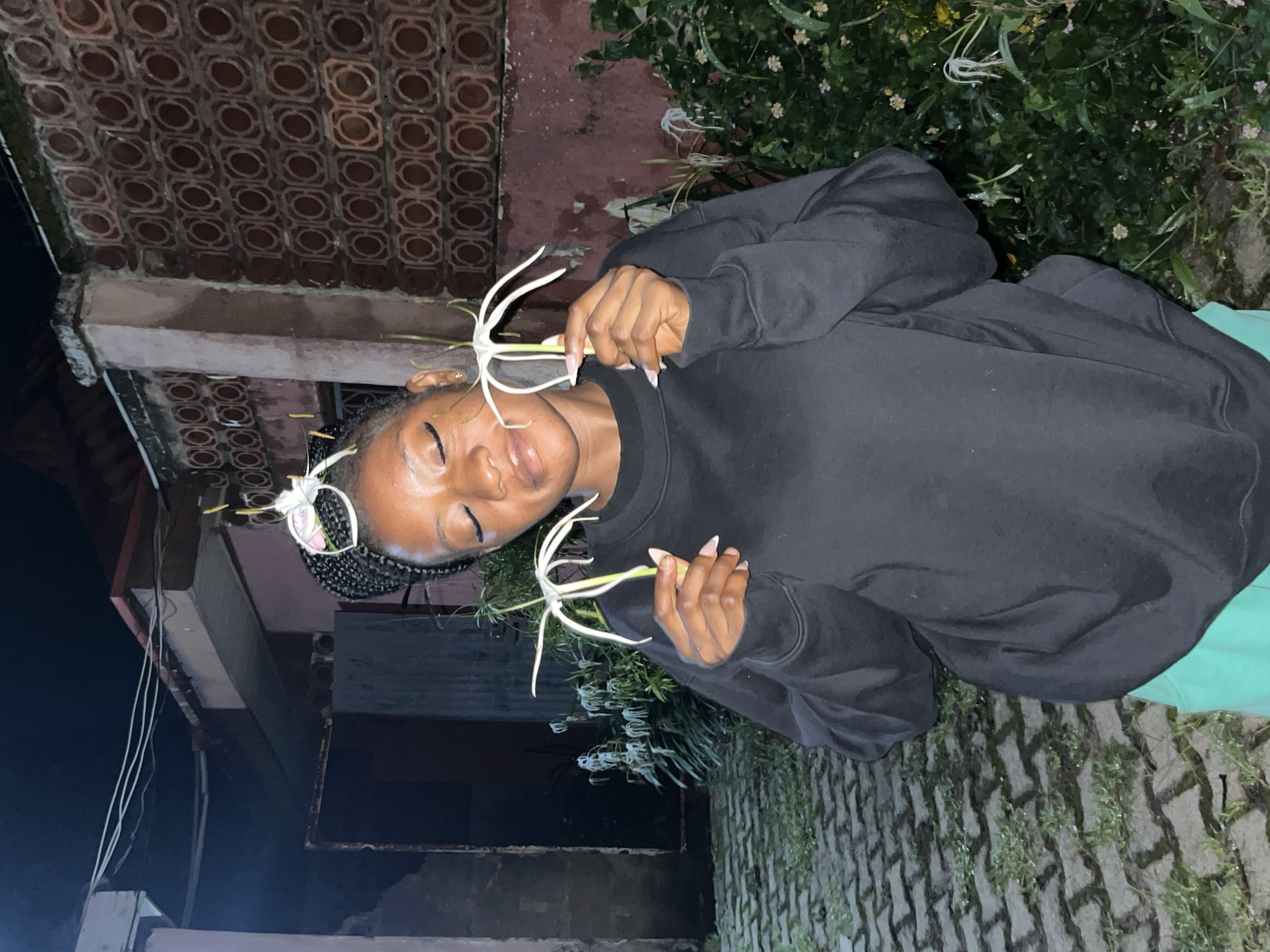 A young Black woman with braided hair is holding two white, spider-like flowers in the city of Jos, Nigeria. She is wearing a black sweatshirt and turquoise shorts. She has her eyes closed and appears to be smelling the flowers. The background shows a brick wall and some greenery. The ground is paved with interlocking stones.