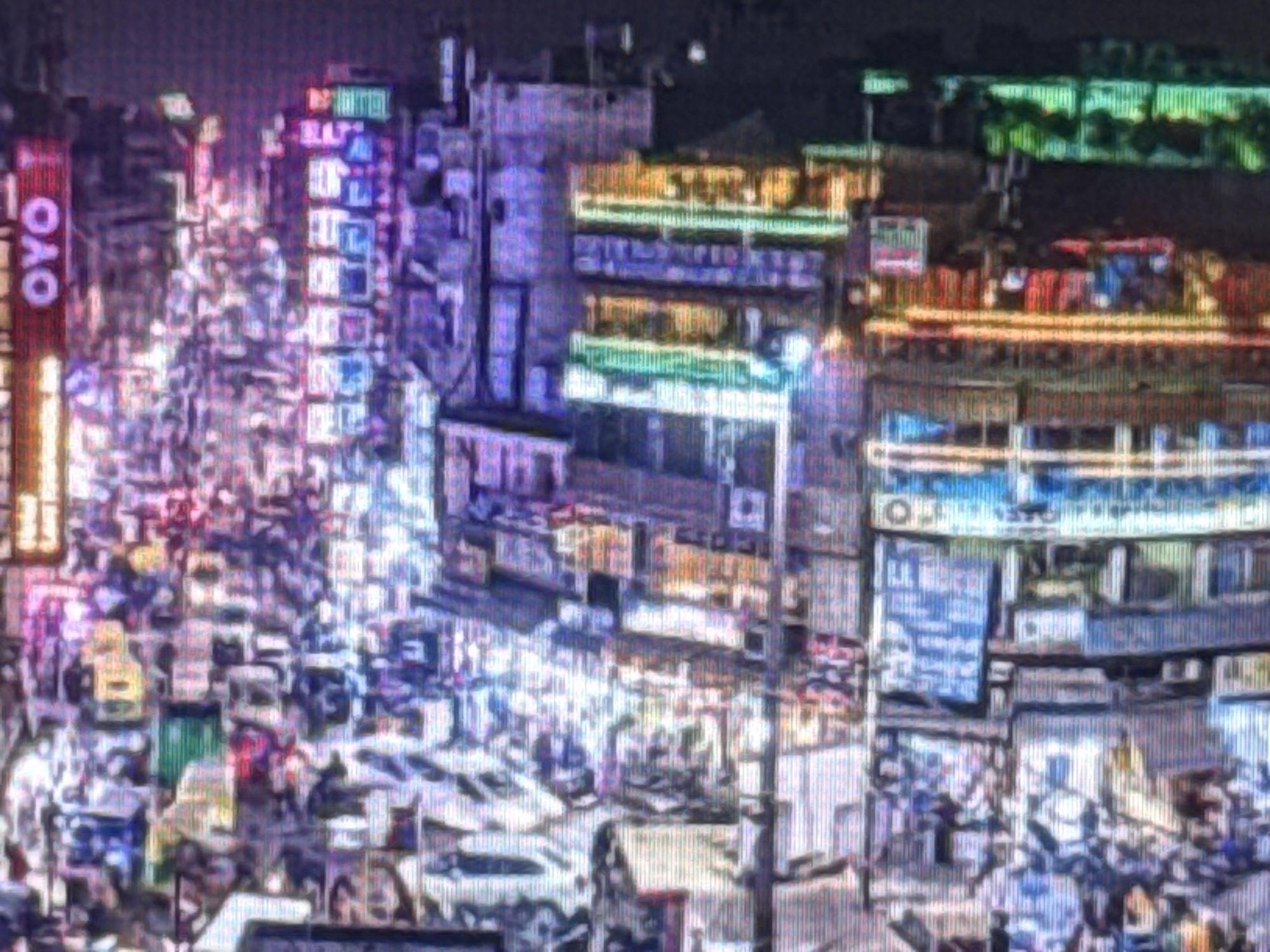 This is a nighttime, low-resolution, pixelated image of a busy city street. The scene is illuminated by numerous neon signs and building lights in various colors, including pink, purple, blue, green, and yellow. The architecture appears to be dense with multi-story buildings lining both sides of the street. There is a significant amount of traffic, with numerous vehicles, including cars and possibly some motorbikes or auto-rickshaws, visible in the street. Some of these vehicles appear to be yellow and white. Pedestrians are also present, though they are largely indistinct due to the low resolution and lighting conditions. The overall impression is of a vibrant and crowded urban environment at night. The specific location is unknown.