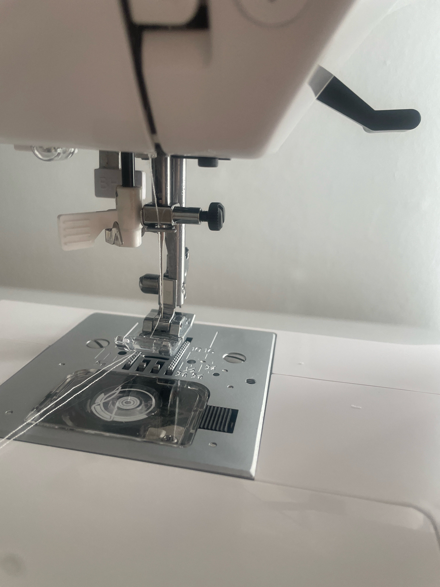 The image shows a close-up view of a sewing machine. The needle is threaded and positioned above the presser foot, which is down on the sewing plate. There is white thread visible, both leading to the needle and on the sewing plate. The sewing machine is white with silver metal accents. Part of a black lever is visible on the right side of the machine. The background is a plain, light-colored wall. The location in Atarfe, Spain, is not depicted or suggested by the image content.