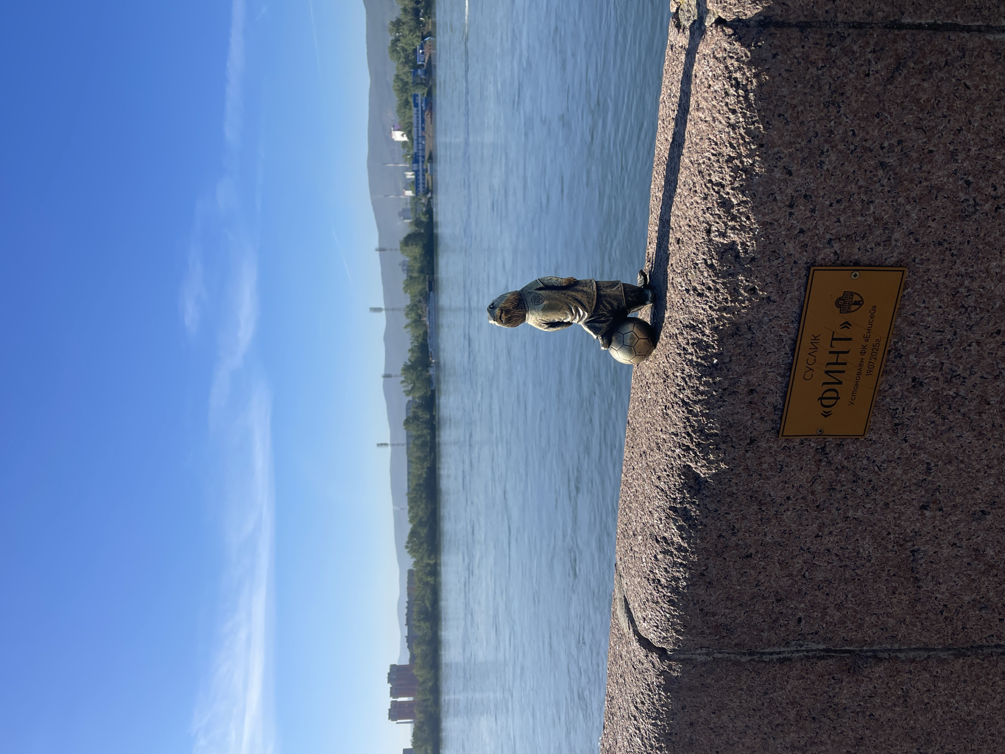 A small bronze statue of a gopher wearing a football jersey and holding a soccer ball stands on a granite embankment overlooking a wide, calm river under a clear blue sky. The statue is positioned to the left of a yellow plaque affixed to the embankment. The plaque contains Russian text, which translates to "Ground Squirrel 'Fint'. Installed by FC Yenisei. 19.07.2025." In the background, across the river, is a city skyline with buildings and industrial structures, backed by distant, hazy mountains. The sun casts a shadow of the statue on the embankment.