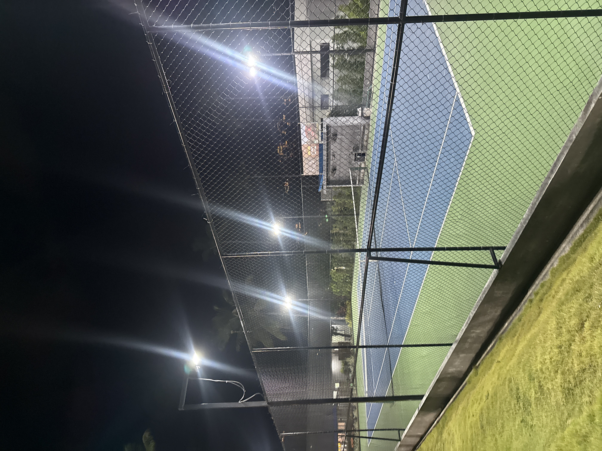 The image shows a well-maintained outdoor tennis court at night, illuminated by bright floodlights. The court is divided into a blue playing surface and a green surrounding area, with white lines marking the boundaries. A chain-link fence encloses the court, and to the left, a strip of manicured grass slopes downwards. In the background, a building with a large graphic display is visible, along with some trees and the distant lights of a city, suggesting an urban or resort setting. The overall impression is of a clean and functional sports facility, likely located in a tropical climate given the lush greenery and palm trees. The specific location of Ko Samui District, Thailand is not directly evident from the image itself, but the environment is consistent with such a locale. There are no people visible in the scene.