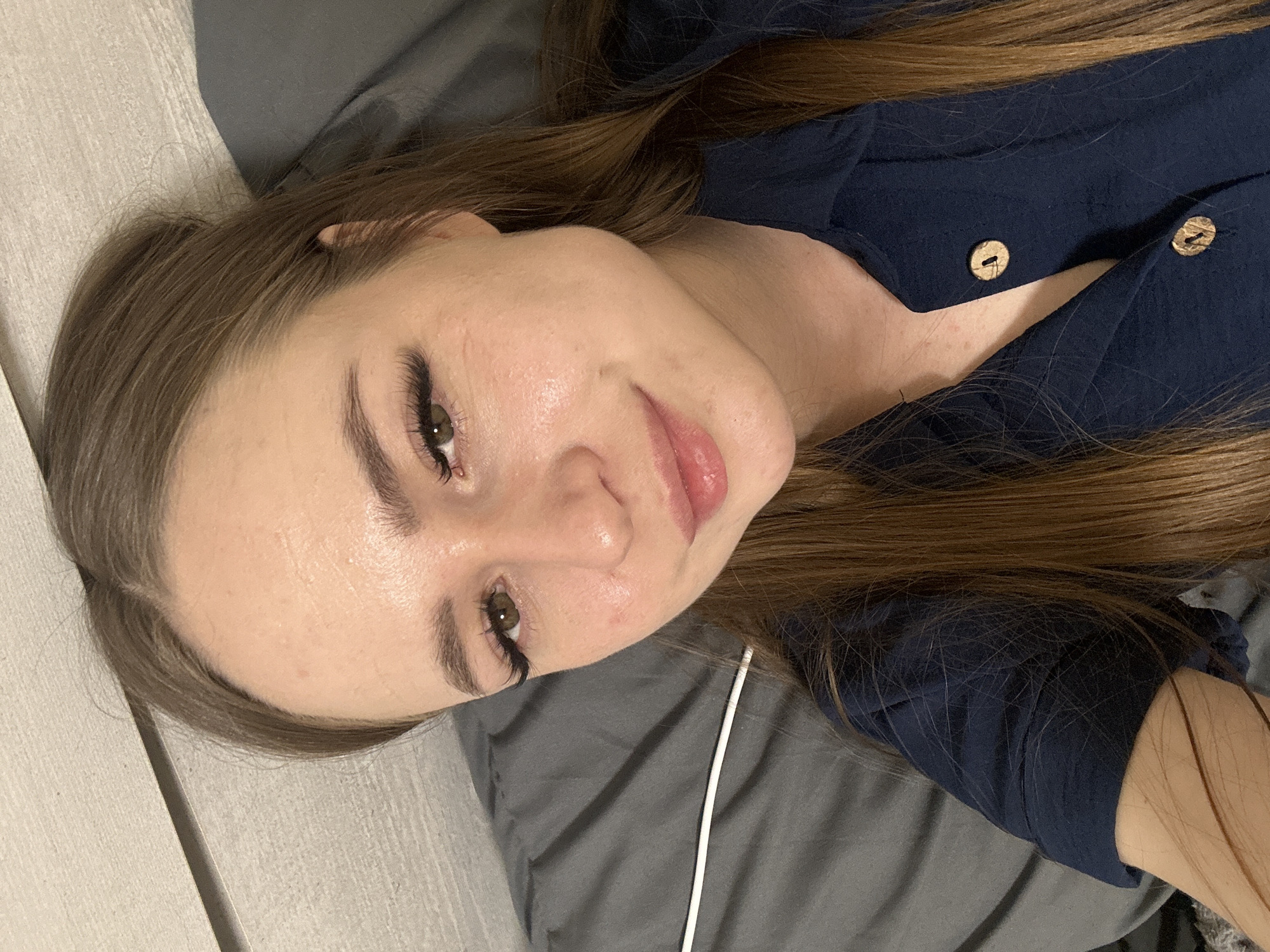 A young woman with brown hair and fair skin is lying on a grey pillow or sheet. She is wearing a dark blue shirt with light brown buttons. Her hair is parted to one side, and she is looking directly at the camera with a slight smile. The background appears to be a bedroom or living space, with a light-colored wall or furniture visible on the left side of the frame. A white cord is visible near her neck.