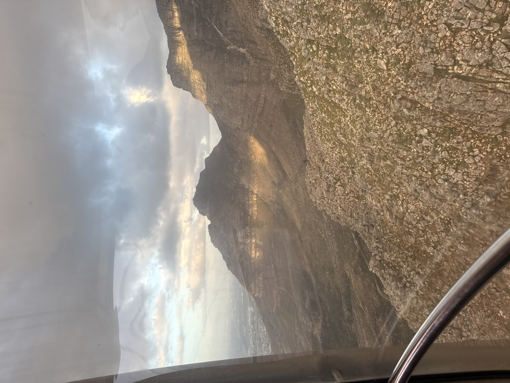 The image shows a view of Table Mountain in Cape Town, South Africa, as seen from a cable car. The landscape includes a rugged mountain face with visible geological layers and scattered vegetation. A city lies in the distant valley, partially obscured by haze. The sky is overcast with clouds. A portion of the cable car's interior is visible, featuring a window frame and a curved metal bar.
