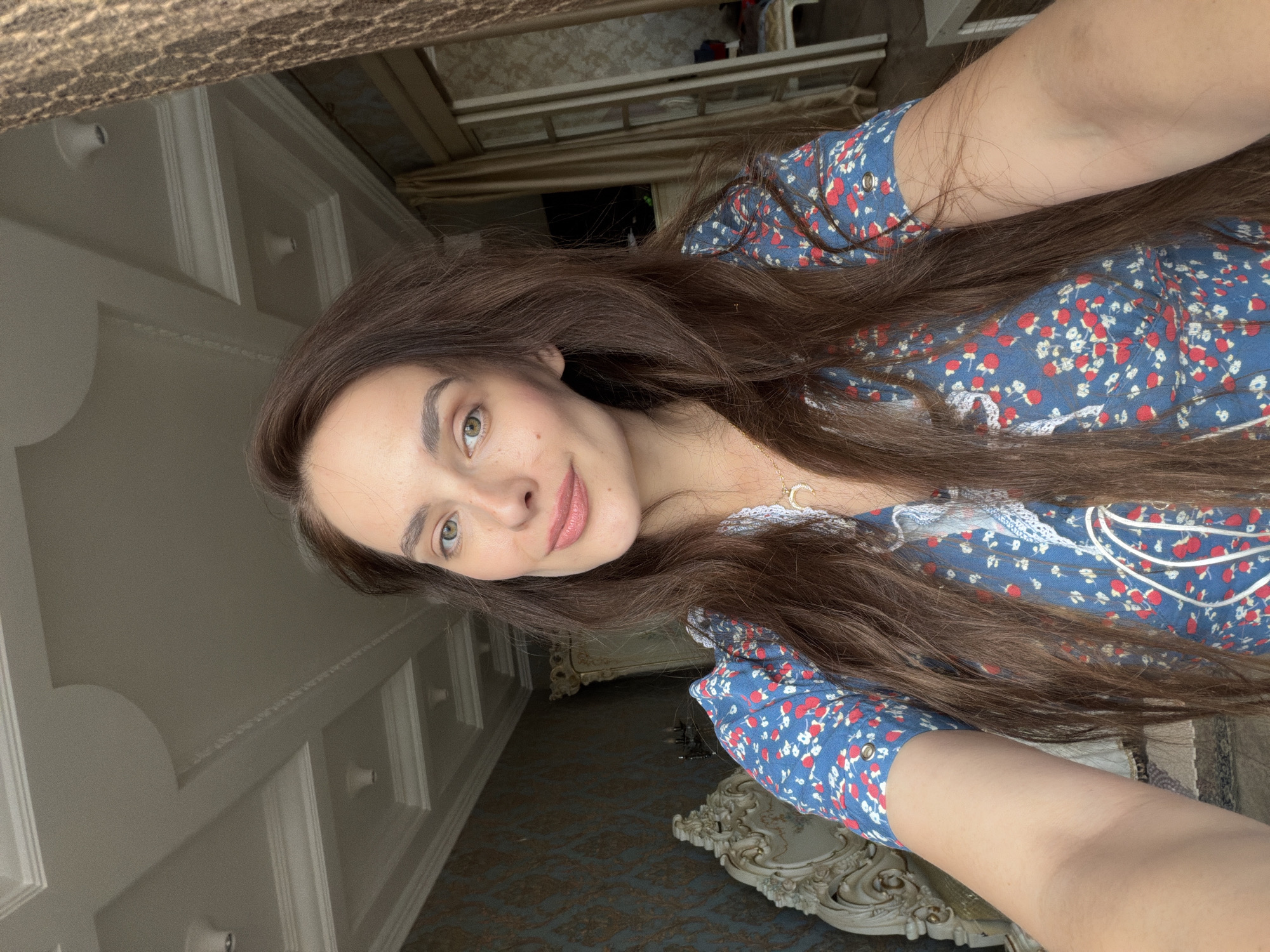 A woman with long, brown hair and green eyes is taking a selfie in a room. She is wearing a blue dress with a floral pattern of red and white flowers. She has a necklace with a crescent moon pendant. The room appears to be a bedroom with ornate furniture, a patterned wallpaper, and a detailed ceiling. There are also curtains and a glimpse of a sliding glass door in the background, suggesting an interior setting, possibly a hotel room or a home. The lighting suggests it is daytime, with natural light entering the room. There is no information to suggest this location is in Ufa, Russia.