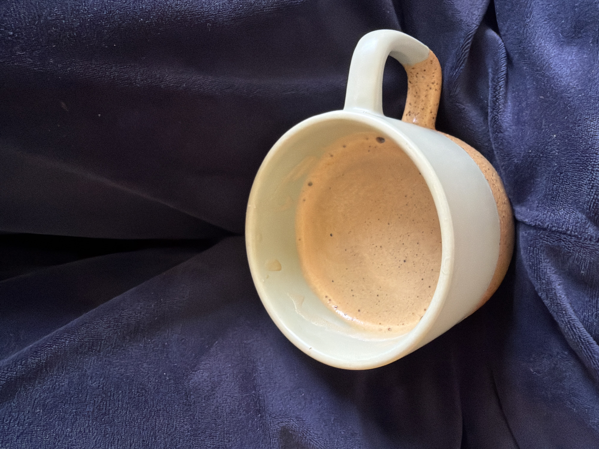 A close-up, high-angle shot features a partially filled, light green and speckled beige mug with a white band and handle resting on a dark blue, possibly velvet, surface. The mug contains a frothy, tan-colored beverage, likely coffee or espresso. The image focuses primarily on the mug and its contents, with the blue surface serving as a background. There is no information to suggest this photo was taken in Pleven, Bulgaria.
