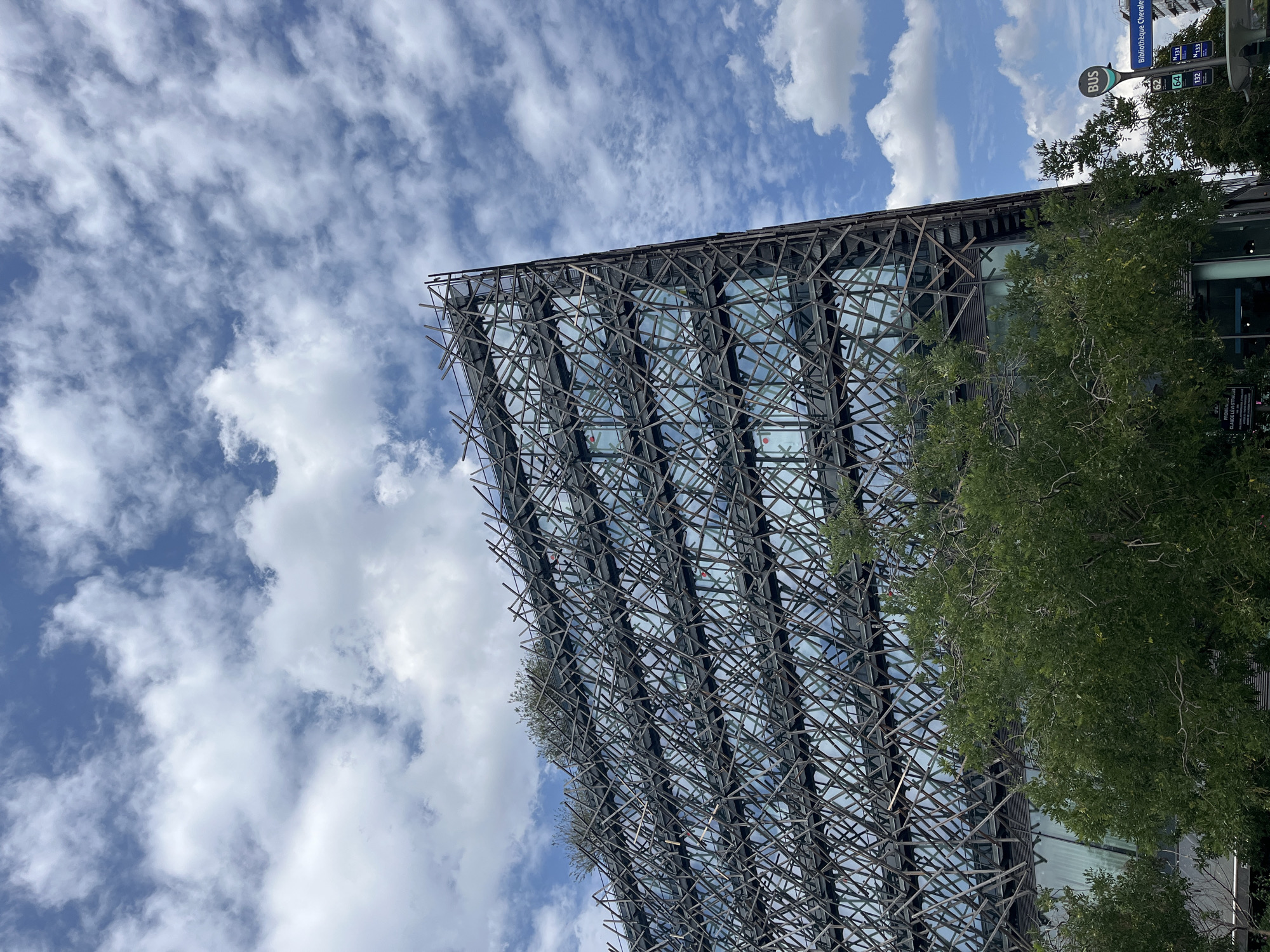 The image shows a modern building with a unique, intricate facade of crisscrossing metal beams. The building appears to be made of glass and metal, with a complex, abstract exterior that resembles a nest or a woven structure. Lush green trees are visible in the foreground and partially obscure parts of the building. The sky above is blue with scattered white clouds, suggesting it's daytime and the weather is fair. There is a bus stop sign in the background, indicating an urban setting. Text visible on the bus stop sign includes "BUS" and route numbers like "62", "64", "131", and "132". Another sign below the bus stop sign appears to have French text, possibly indicating the location as "Bibliothèque Chevaleret". The perspective is from below, looking up at the building and the sky. There are no people visible in the frame. The overall impression is one of striking contemporary architecture set against a natural element and a typical urban feature.