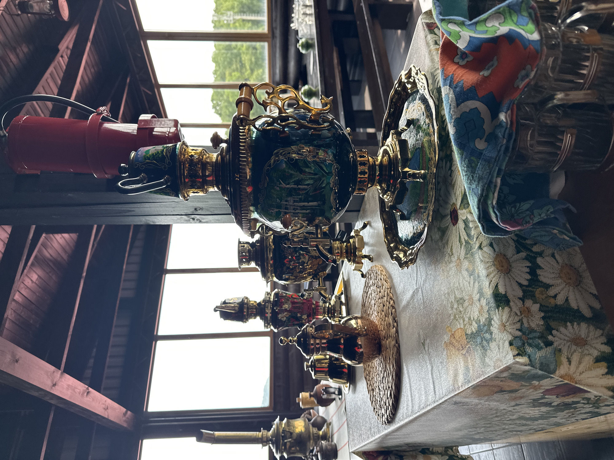 An indoor setting, possibly a cafe or restaurant in Sochi, Russia, features a collection of ornate samovars and teapots arranged on a tablecloth adorned with a floral pattern of daisies. The room has a rustic ambiance with exposed wooden beams supporting the ceiling and large windows offering a view of greenery outside. In the foreground, a large, dark green samovar with gold accents is prominent, resting on a gold tray. Beside it, a smaller, mirrored teapot sits on a woven placemat. Various other samovars and teapots, some appearing to be brass and others with decorative paintings, are scattered across the table. A colorful patterned cloth is draped over what appear to be glass mugs in the lower right corner. In the background, wooden tables and benches are visible, along with more glassware, suggesting a dining or gathering area. A red apparatus hangs from the ceiling above the main samovar. The lighting suggests it is daytime.