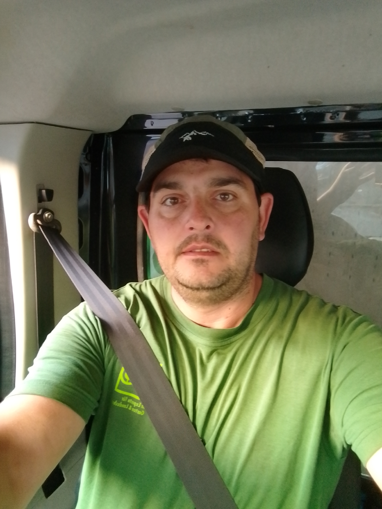 A man in a green t-shirt and a black and tan baseball cap is seated in a vehicle, likely a van, as indicated by the interior paneling and seatbelt visible. He is wearing a dark gray seatbelt across his chest. His expression is neutral, and he is looking directly at the camera. The background shows the interior of the vehicle, with a dark headrest and a window reflecting an outdoor scene. The lighting suggests it is daytime. The image appears to be a selfie taken from the driver's seat. The location is identified as Wedel, Germany.