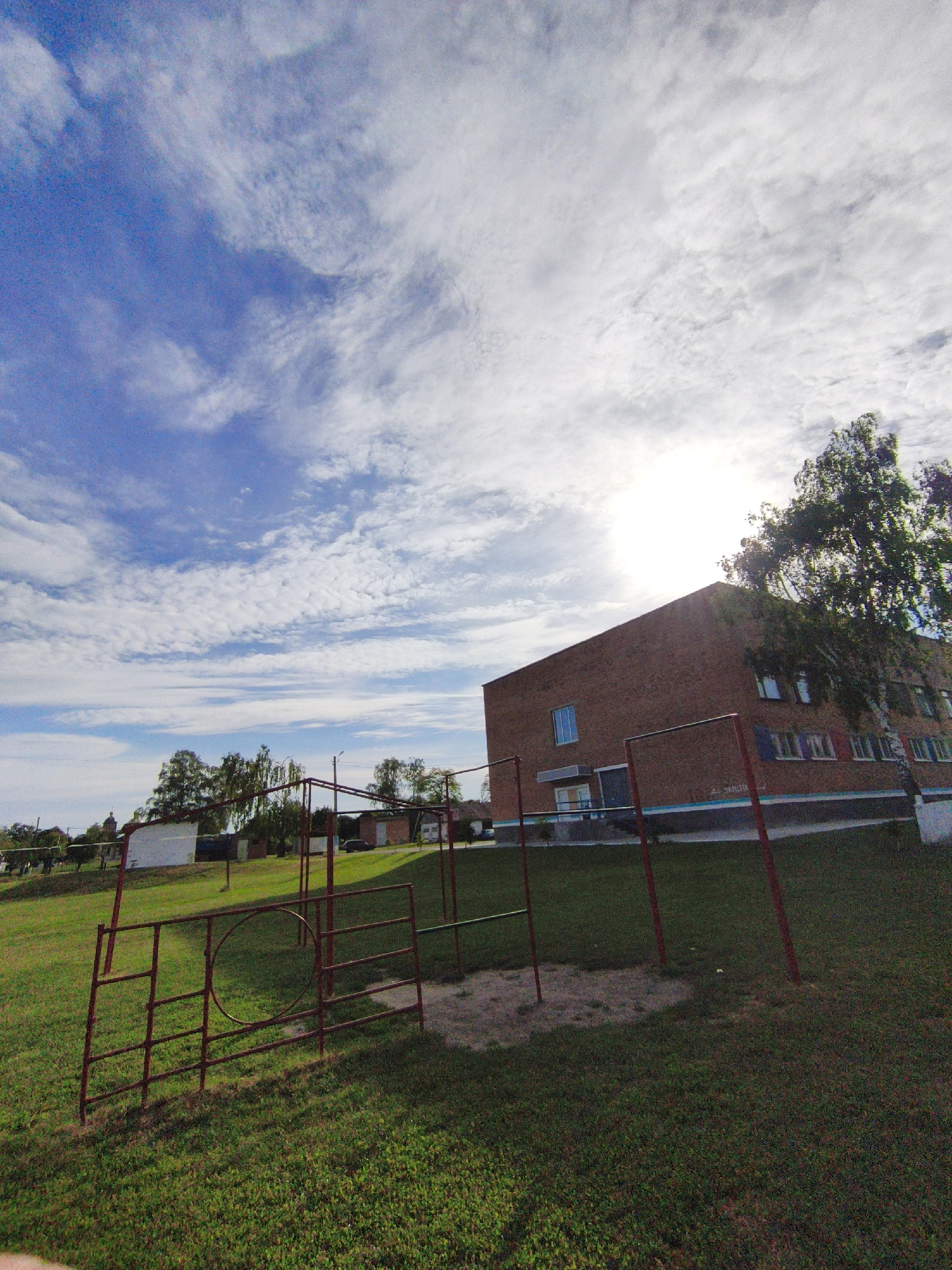 The image captures an outdoor scene, likely a schoolyard or public space in Ustyvytsia, Ukraine, under a bright, partly cloudy sky. The sun is positioned high on the right, creating a bright glare and casting shadows across the foreground. In the immediate foreground and midground, a green grassy area features red metal gymnastic or playground equipment, including parallel bars and a climbing frame with a distinctive circular element, set over a patch of sand. To the right, a large, multi-story brick building, possibly a school, is prominent, with several windows and a tall, slender tree standing next to it. Faint Cyrillic text, reading "ДО ШКОЛИ" (To School), is visible on a blue-painted section of the building's facade. The background on the left reveals distant buildings and trees, with the subtle outline of a church dome further away. The scene is devoid of people, conveying a peaceful and static moment during daylight hours.