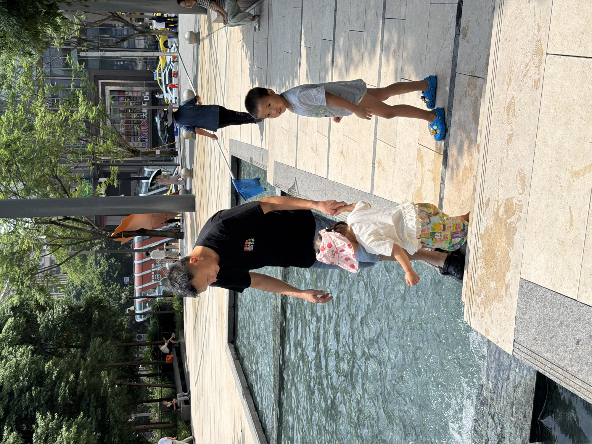 In Yuzhong District, China, on a sunny day, a man in a black t-shirt and denim shorts stands by a water feature with his right hand outstretched towards the water. Two children are near him. A young girl in a white shirt and a pink sun hat with strawberries on it, wearing colorful shorts, is facing away from the camera, looking at the water. A young boy in a grey t-shirt and blue Crocs is facing the camera, looking to his right. The man is holding the girl's hand. In the background, other people are visible, including a person in a blue shirt walking with a long pole and a net. The environment appears to be a public plaza with trees, buildings, and vehicles in the distance.