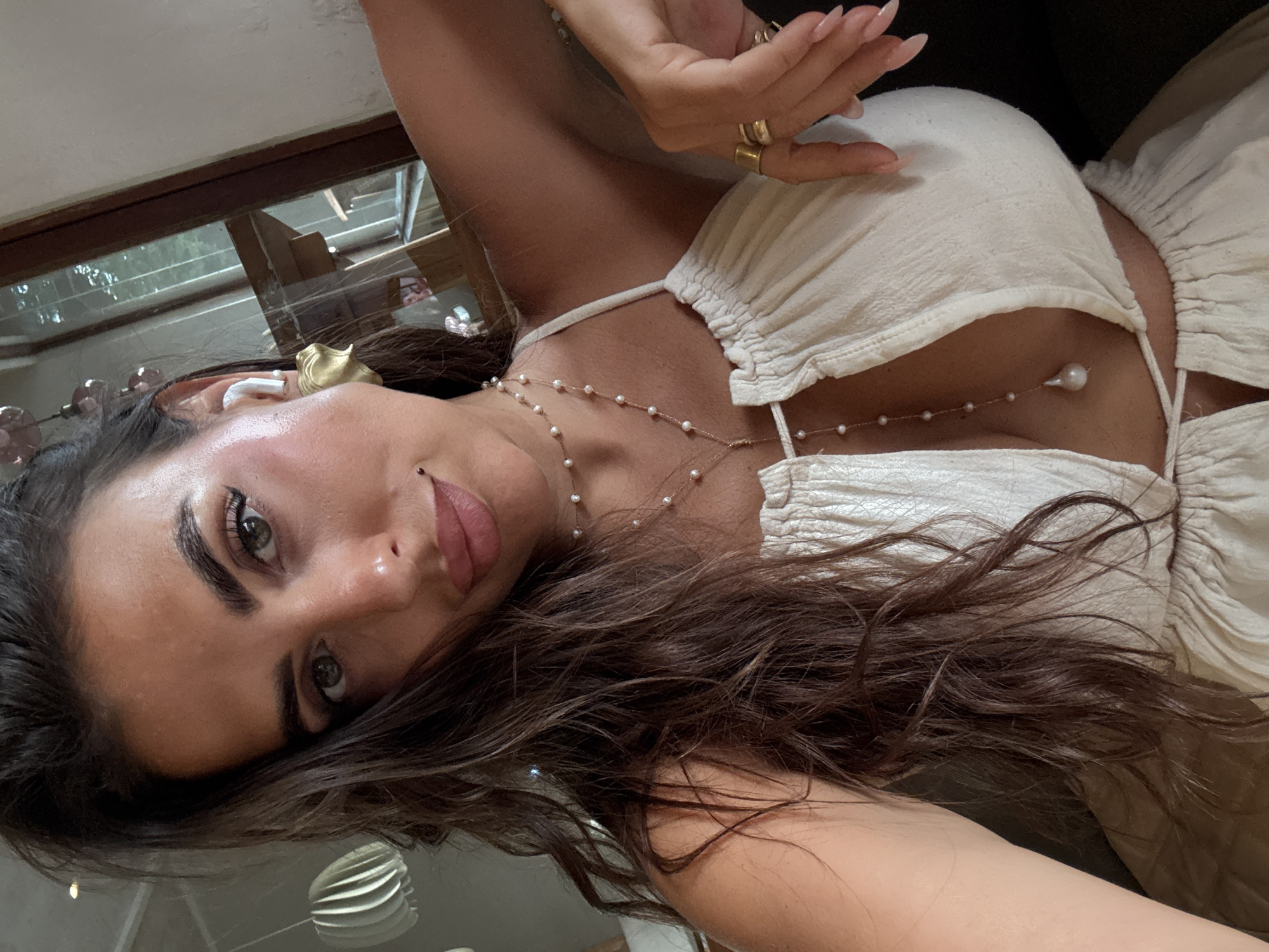 A woman with dark hair and fair skin is photographed from a low-angle perspective. She wears a white top with a pearl necklace. She has a small piercing on her upper lip. She wears an in-ear wireless device and a gold-colored earring. She has several gold rings on her fingers. Her location is North Kuta, Indonesia.