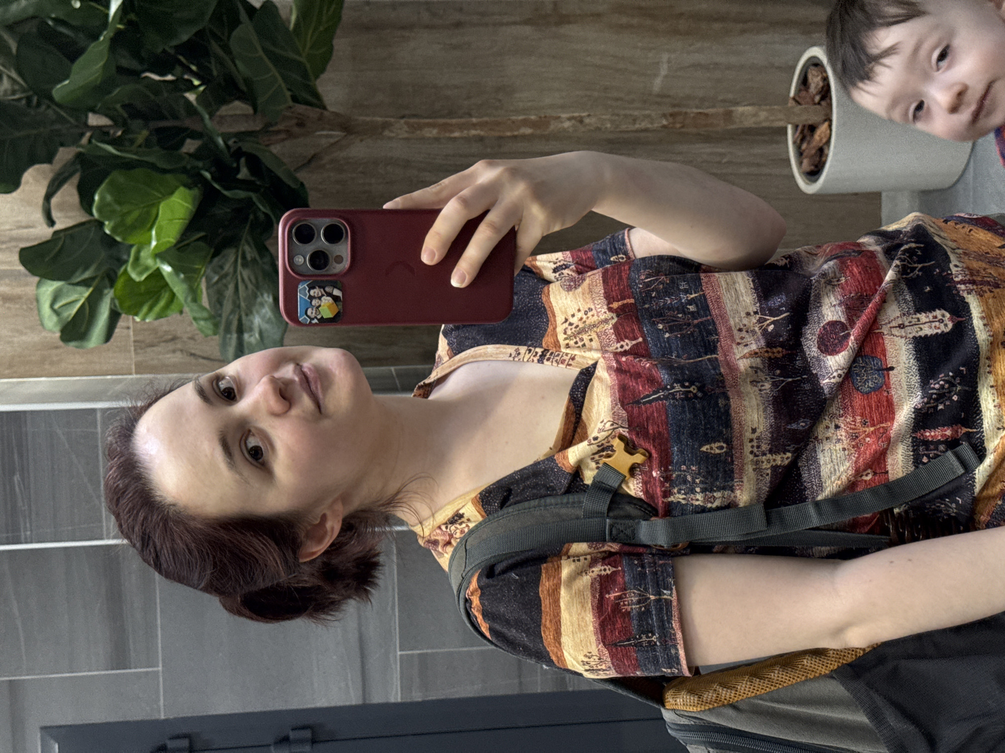 A woman is taking a selfie with a red phone. She is wearing a patterned top and a backpack. A child is visible in the upper right corner. A potted plant stands in the background. The scene features tiled walls and a wooden surface. There is no information to determine if the location is in Izhevsk, Russia.