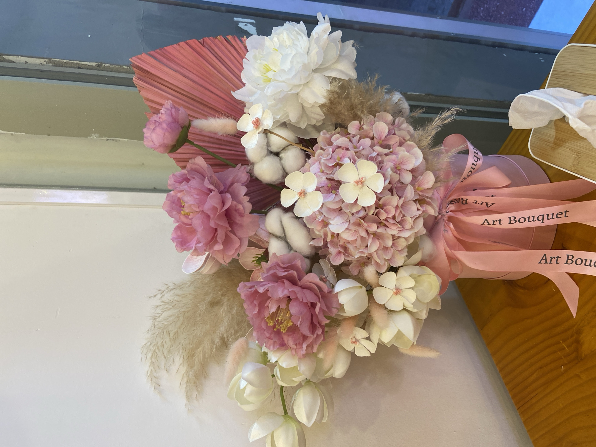 This media file displays a close-up, top-down view of an artificial flower arrangement. The bouquet features a variety of flowers in shades of pink, white, and cream, including peonies, hydrangeas, and tulips. Pampas grass and cotton bolls are also incorporated into the arrangement, adding texture and visual interest. A pink fan-shaped palm leaf serves as a backdrop. The bouquet is tied with a pink ribbon that has the words "Art Bouquet" printed on it in dark text. The bouquet appears to be placed in or near a cylindrical pink container, also adorned with the same ribbon.
The background suggests an indoor setting. To the left, a window with a blue frame and a white sill is visible, hinting at natural light entering the space. The windowpane reflects a dimly lit area. To the right, a wooden surface, possibly a table or shelf, is visible. On this surface rests a light-colored wooden box and a crumpled white tissue or paper. The overall impression is one of a decorative item displayed indoors, likely in a home or shop. There are no people present in the frame. The lighting suggests it is daytime, but the specific time is not discernible. The scene is static, with no activity taking place. The focus is entirely on the arrangement of artificial flowers. The visible text on the ribbon clearly states "Art Bouquet".