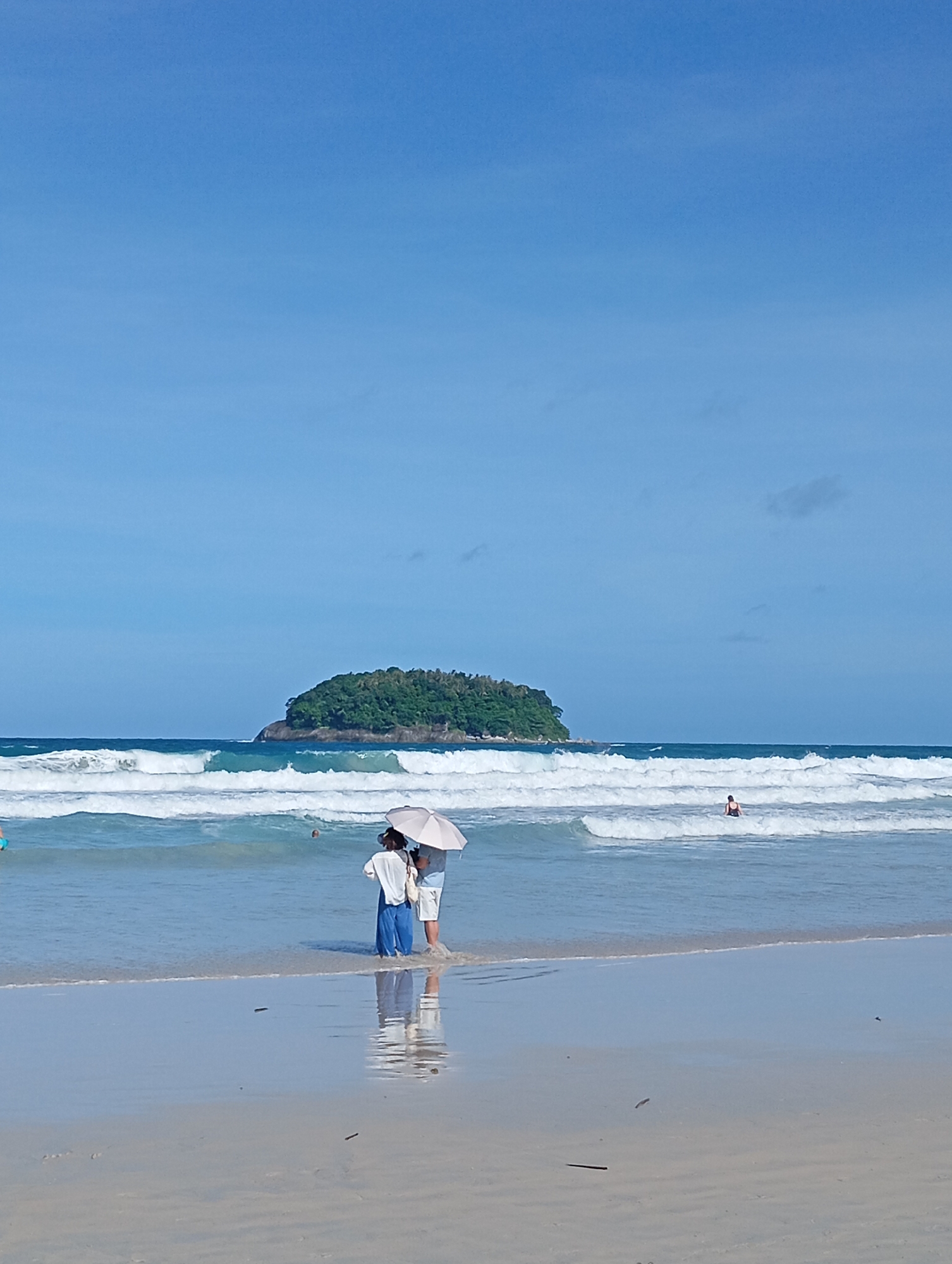 A wide-angle, eye-level shot captures a bright, sunny day on Karon Beach, Thailand. The scene is dominated by the vast, clear blue sky and the shimmering turquoise ocean. In the middle ground, gentle white-capped waves roll onto the sandy shore. A small, lush green island sits on the horizon, adding a touch of tropical beauty to the seascape.
In the foreground, two individuals stand near the water's edge, sharing a light-colored umbrella. One person, wearing a white top and blue pants, is partially obscured. The other, in white shorts and a white top, stands beside them. Their reflections are visible in the shallow water. Further out in the ocean, a person in a dark swimsuit is visible wading in the waves. A sliver of the sandy beach is present in the lower portion of the frame. The overall impression is one of a peaceful and serene beach day.