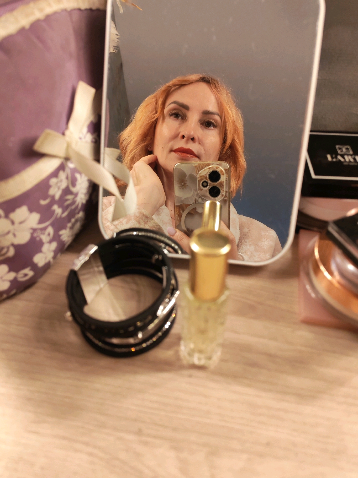 A woman with short, orange hair is taking a mirror selfie. She is wearing makeup, including red lipstick. In the foreground, there is a black bracelet with rhinestones, a small glass perfume bottle with a gold cap, and a portion of a purple fabric item with a white ribbon. In the reflection, a black and gold box with the word "EARTH" is visible. The setting appears to be a dresser or vanity. The image was taken in Chaikovsky, Russia.