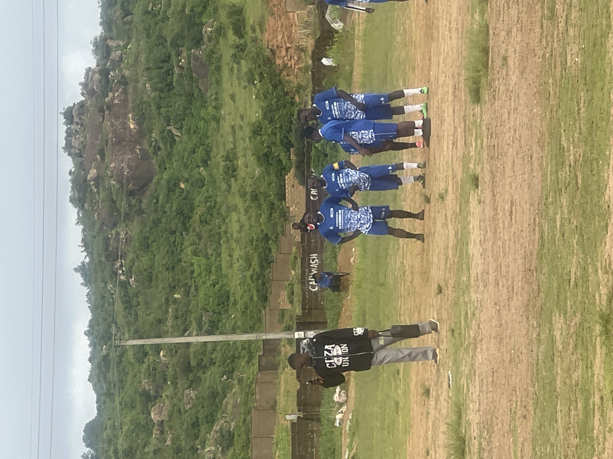 This image shows a group of young men in blue and white soccer uniforms standing on a dusty field with sparse grass. They appear to be a team, possibly a local club, given the uniforms and the informal setting. In the background, a man in a black hoodie and gray pants is looking at his phone, holding a black bag. There is a brick wall with graffiti that reads "CARWASH" and further back, a rocky, tree-covered hillside typical of the landscape around Jos, Nigeria. A utility pole stands near the center of the frame. The sky is overcast, suggesting a cloudy day.