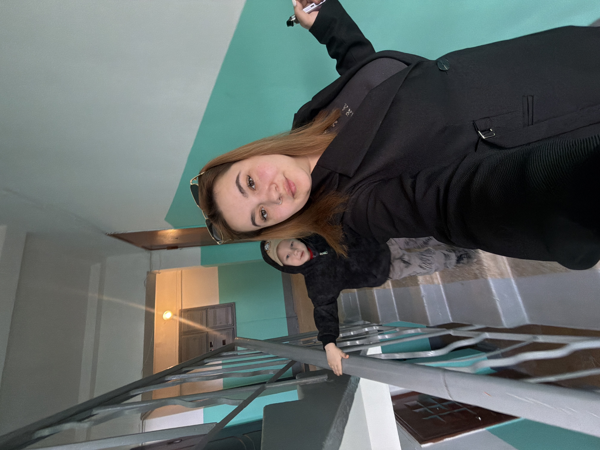 Location: Apartment building staircase in Chaikovsky, Russia.
Scene: A woman is taking a selfie while standing on a staircase. She is wearing a black coat and has long brown hair. A young child in a black hooded jacket and a light-colored hat is visible behind her, holding onto the railing. The staircase has metal railings and concrete steps with colored accents. The walls are painted with white, blue, and green sections. A light fixture is visible in the background, illuminating a doorway.
