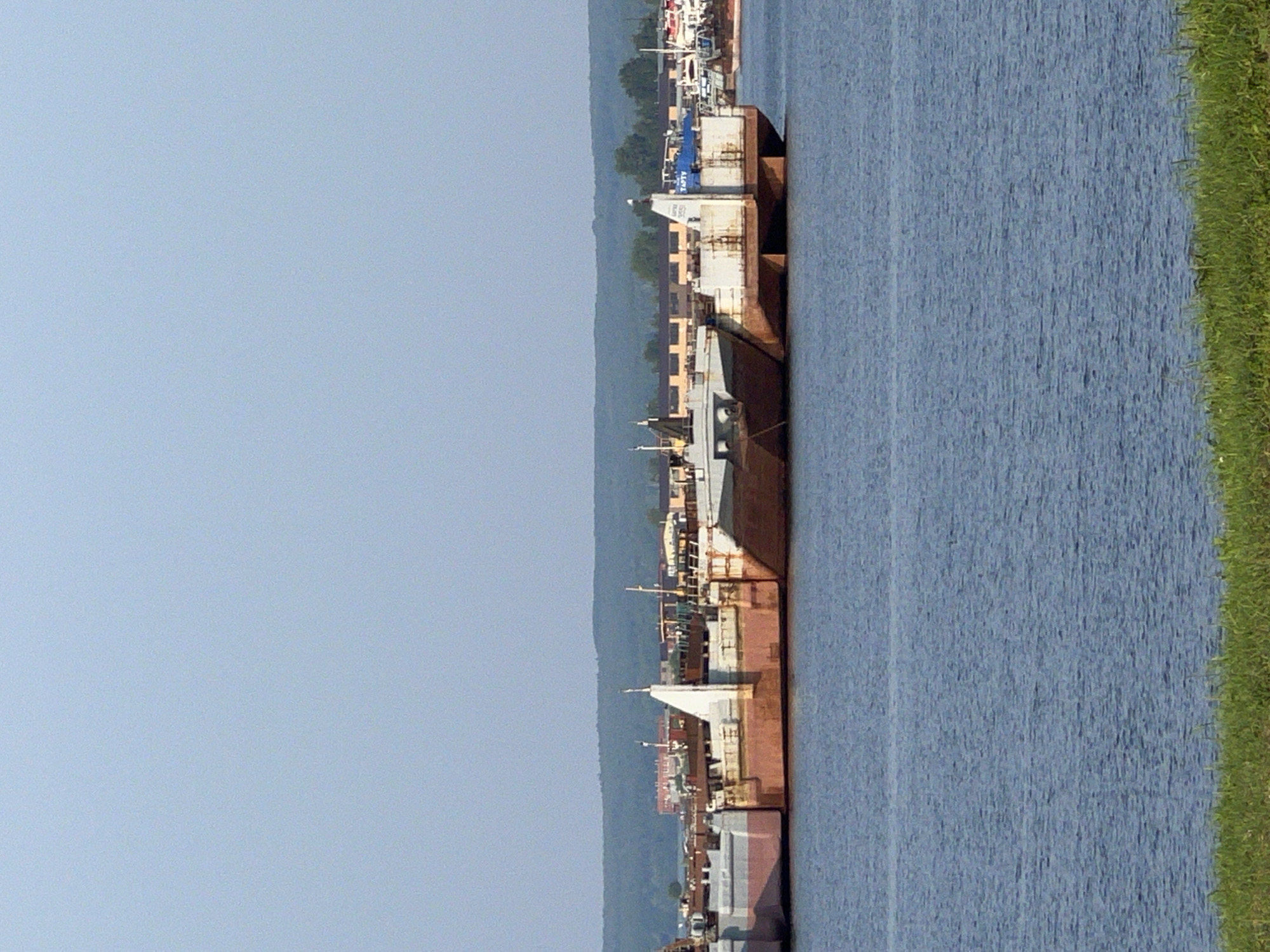 A row of decommissioned or moored barges are lined up in the Kama River near the city of Chaikovsky, Russia. The barges are primarily rusted and weathered, with some sections showing red and grey paint. They are of various sizes and designs, some appearing to be flat-bottomed cargo barges. In the background, there is a distant shoreline with a line of trees and some buildings, including what appears to be a brick structure and some industrial-looking buildings. The sky is clear and pale blue. The foreground shows the calm, blue water of the river and a strip of green grass along the bank. There are no people visible in the image.