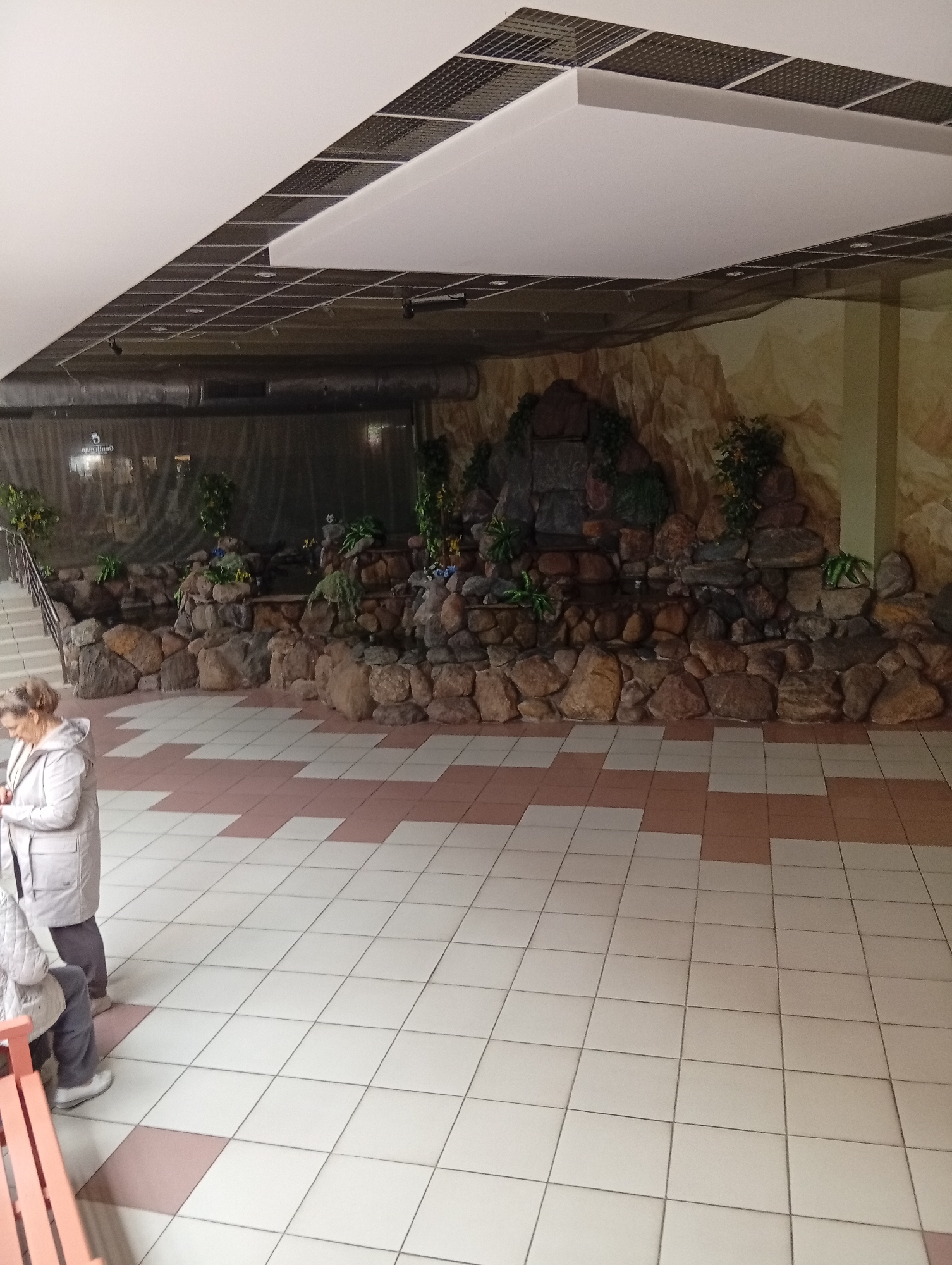 This image captures an indoor scene featuring a decorative rock waterfall with artificial greenery against a mural of a mountainous landscape. A tiled floor, patterned with white and pinkish-brown squares, extends into the foreground. In the left portion of the image, a woman wearing a grey hooded jacket and dark pants stands looking down. The ceiling is a drop ceiling with recessed lighting and what appears to be acoustic paneling. The overall impression is that of an interior public space, possibly a shopping center or an atrium. The location in Šiauliai, Lithuania is not discernible from the visual information provided in the image.