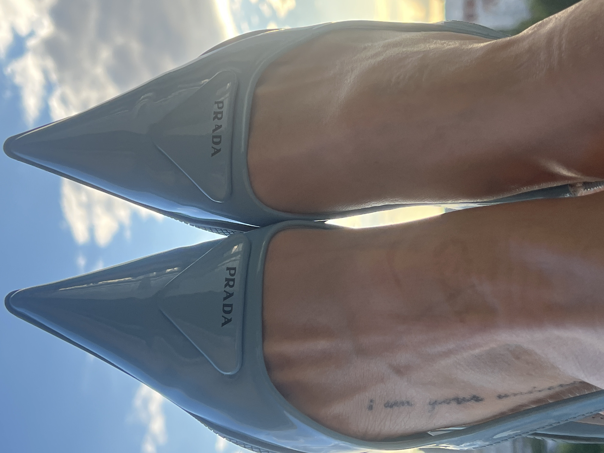 A close-up, low-angle shot shows a pair of pale blue, patent leather Prada stiletto heels, worn by an unseen person. The shoes are sharp and pointed, with the Prada logo subtly embossed on a triangular detail near the heel. The focus is on the toes of the shoes and the wearer's ankles and feet, which are exposed. A small, vertical tattoo is visible on the left ankle. The background consists of a bright, cloudy sky, suggesting an outdoor setting. The image does not provide any information about the specific location in Saint Petersburg, Russia.