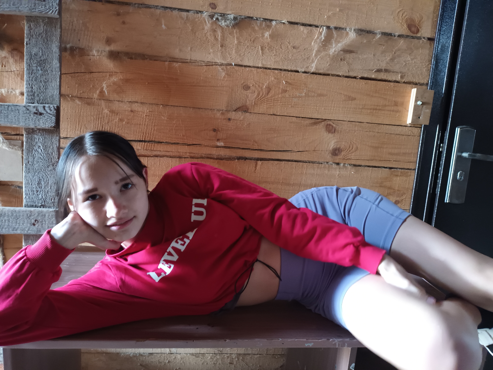 A young woman is lying on a wooden surface in what appears to be a cabin or rustic room. She is wearing a red cropped sweatshirt with white lettering and lilac-colored exercise shorts. The background consists of wooden planks that form the wall and a black doorframe is visible to the right. The location is Ольховка, Russia.