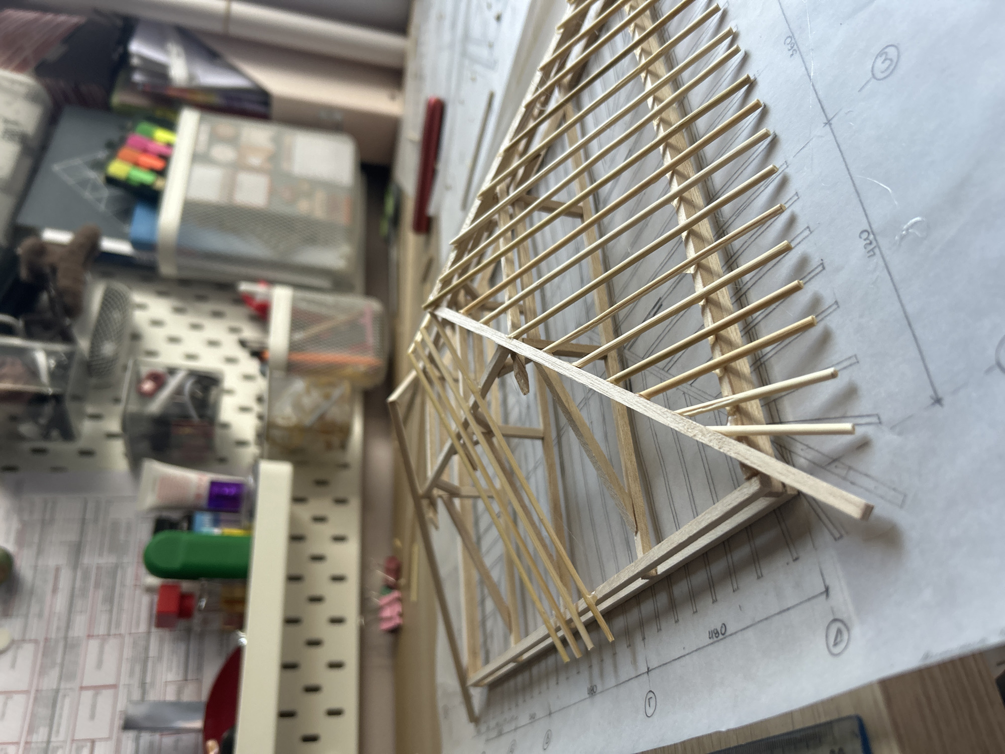 The image displays an architectural model crafted from light-colored wooden strips, positioned on top of a technical drawing or blueprint. The model, prominently featured in the right and center, appears to represent a roof structure or a complex framework of a building, characterized by numerous parallel and intersecting wooden elements forming a skeletal design.
Beneath the model, the white blueprint paper shows black lines, measurements, and symbols. Visible text on the blueprint includes numbers such as "560", "420", and "480", along with circled alphanumeric identifiers like "3", "D", and "Γ".
The background to the left is blurred, revealing parts of a workspace. A white pegboard system is visible, holding various office supplies and organizers. Partially discernable items include colorful highlighter pens in a holder, a small white desk fan, and other containers for tools or stationery. The scene suggests an indoor environment, likely an architect's studio, a design workshop, or a student's desk, where an architectural project is being developed or assembled. The activity is centered around the creation or study of this scale model in relation to its technical plans. The location context is Sofia, Bulgaria, though no specific visual elements in the image confirm this geographical detail.