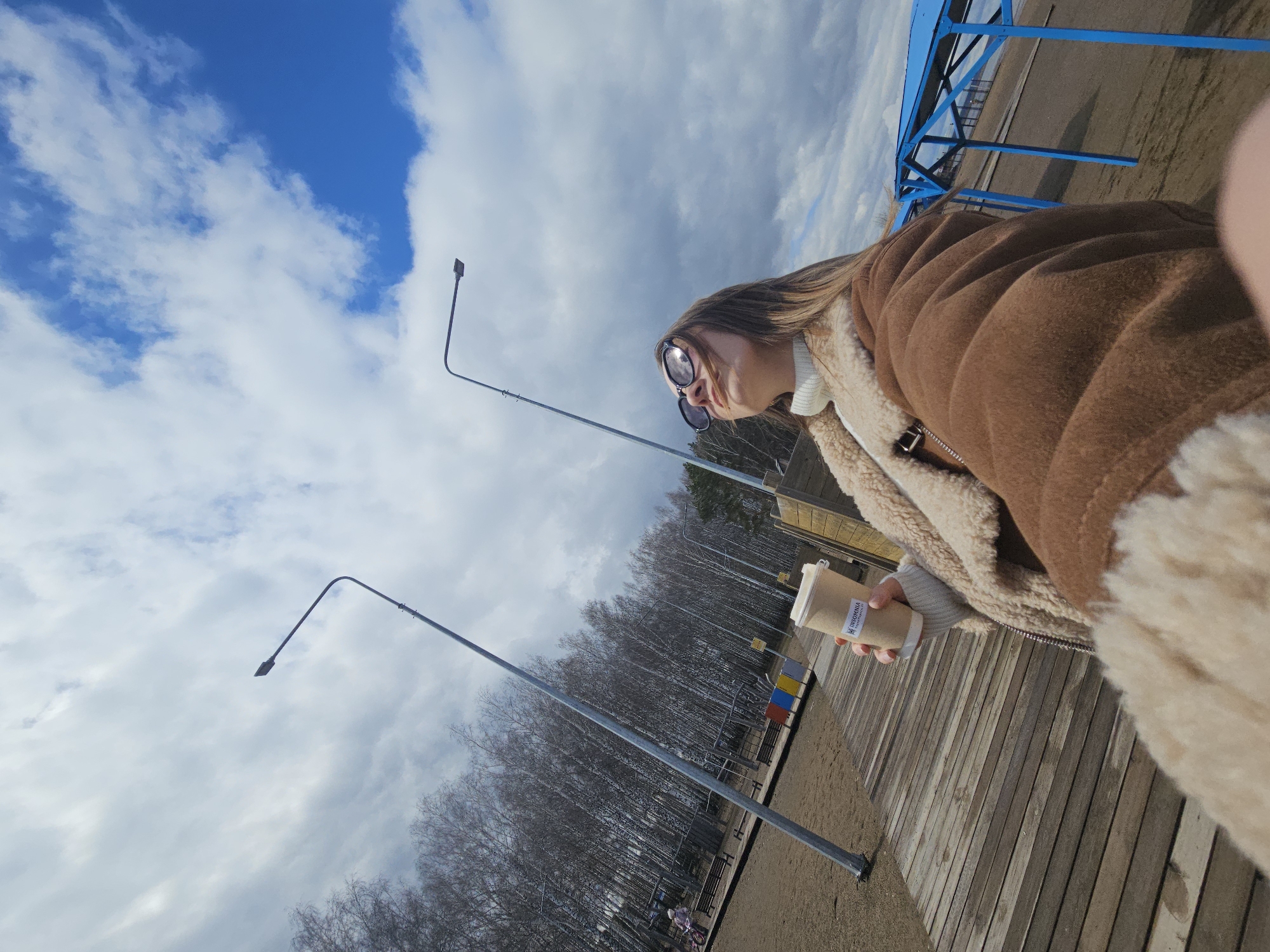 A young woman is seen in an outdoor setting, potentially the city of Chaikovsky, Russia. She is wearing a brown jacket with a white sweater and sunglasses. The subject is holding a beverage in her hand. In the background, there is a line of bare trees, street lights, a wooden deck, and a blue-colored structure. The sky contains a mix of blue and white clouds.
