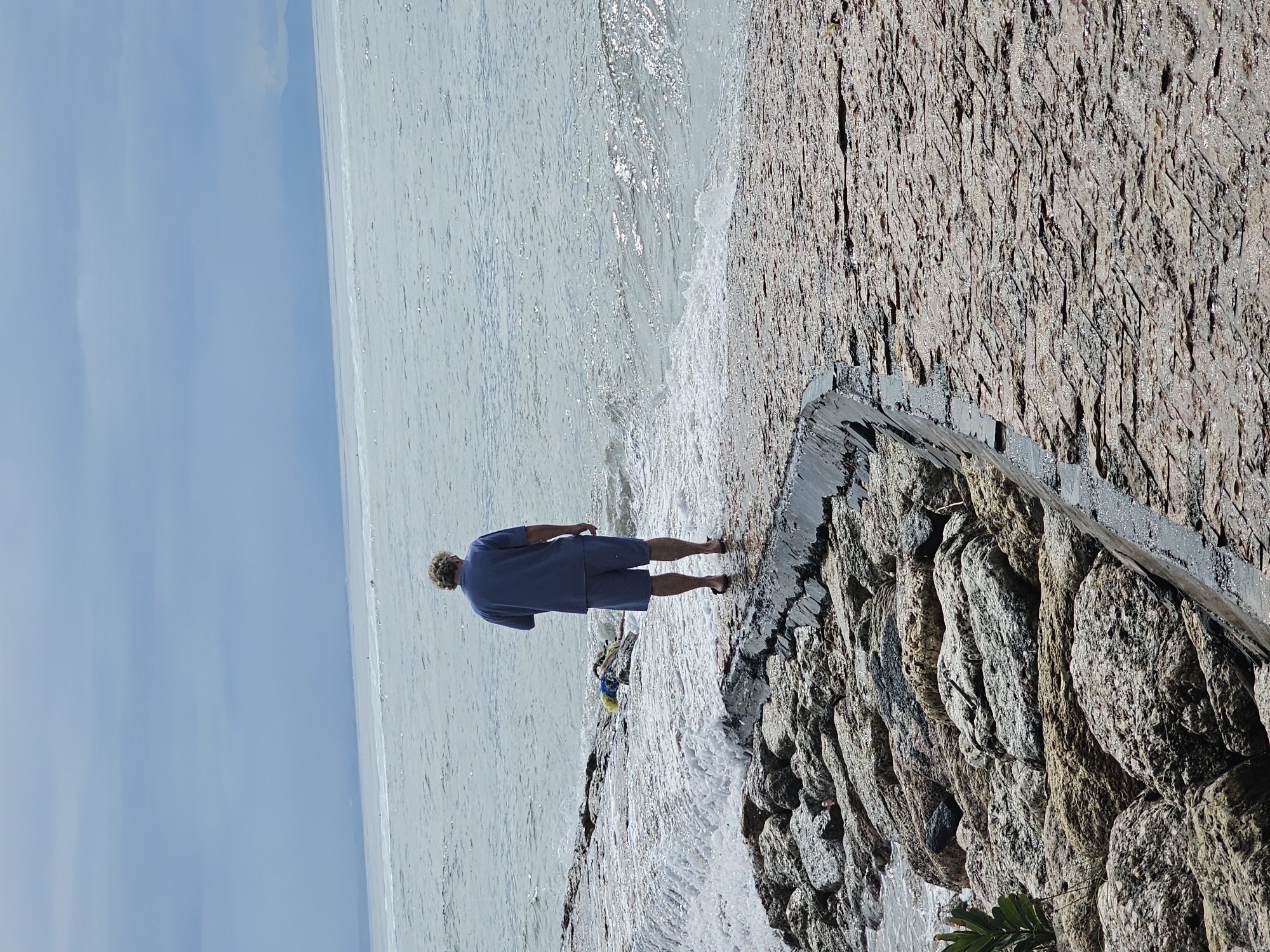 A man stands facing the sea, near a stone wall, in Denpasar, Indonesia. The man is wearing a blue shirt and shorts. Waves are crashing against the wall. The sky is pale blue.