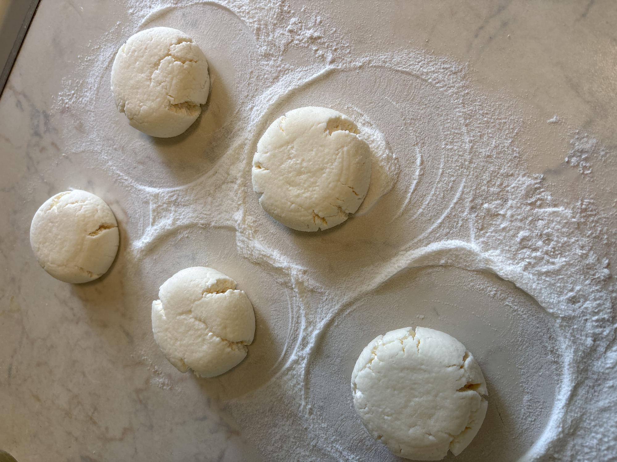 Five white, disc-shaped objects are arranged on a marble surface dusted with flour. Each object appears to be a dough or batter preparation, with visible cracks and a slightly uneven texture. They are scattered across the surface in a somewhat haphazard pattern, with flour creating subtle outlines around some of them. The lighting suggests an overhead source, casting soft shadows. This scene is located on a countertop in Rusanovo, Russia.