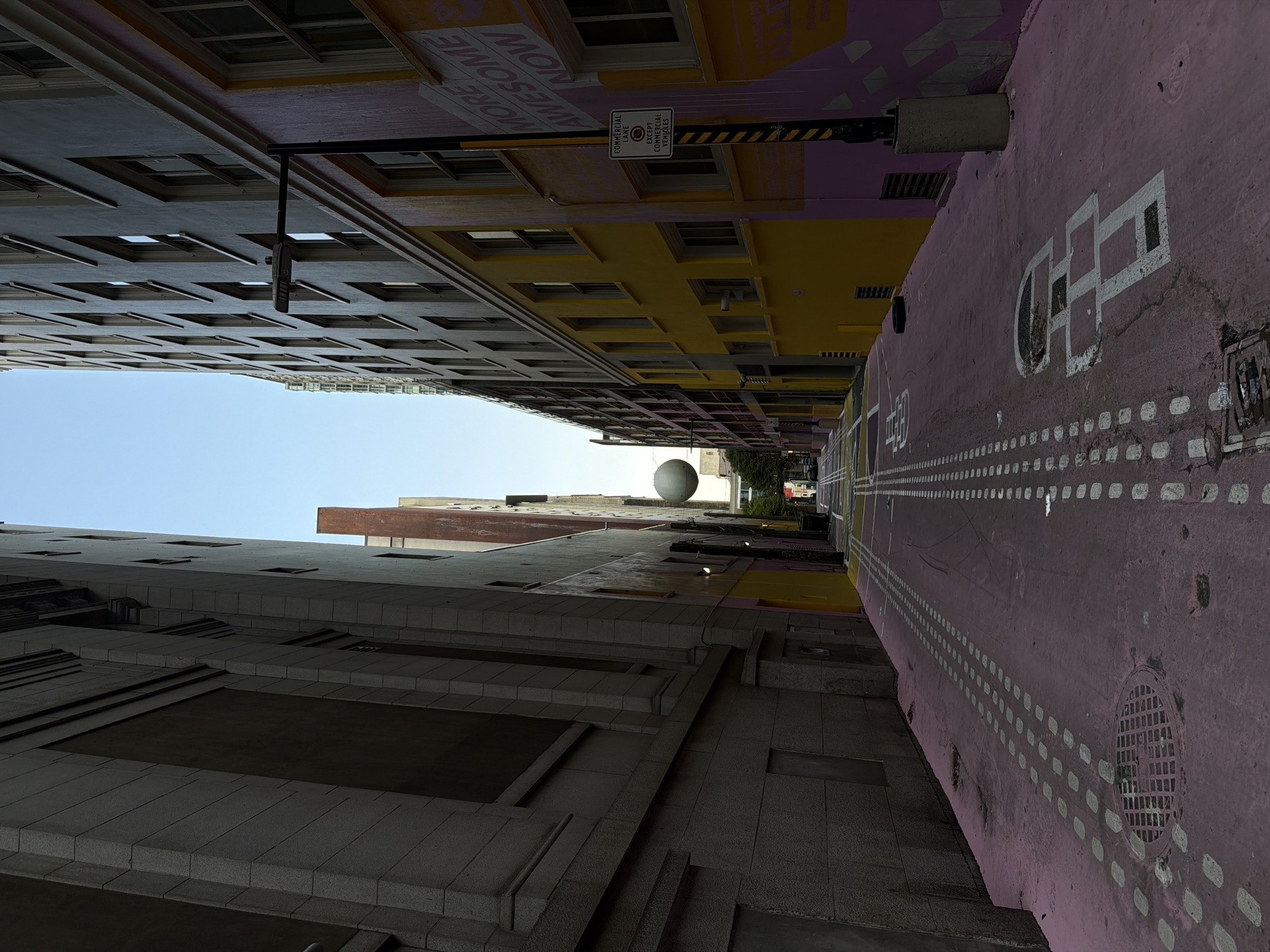 The image depicts a narrow city alleyway in Vancouver, Canada, which has been painted pink with white dotted lines and hopscotch diagrams on the pavement. Tall buildings line both sides of the alley, with the building on the left appearing to be made of grey stone and the building on the right painted in bright yellow and pink with graffiti that reads "MORE AWESOME NOW". A street light is mounted on the right-hand building. In the distance, a large white spherical sculpture is visible suspended in the air. The sky is clear and blue. The alley appears to be empty of people and vehicles, suggesting a quiet moment in an urban setting.