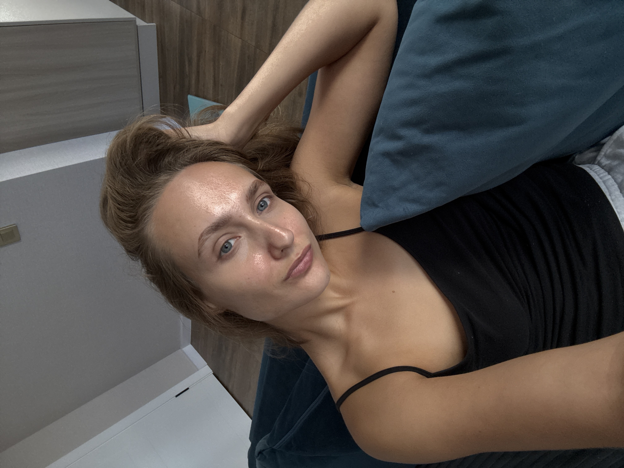 A young woman with blonde hair and blue eyes is lying on a teal couch, her head resting on a teal pillow. She is wearing a black tank top. Her left arm is bent, with her hand resting on her head. The background shows a wooden floor and a white door. The image appears to be taken indoors in an apartment.