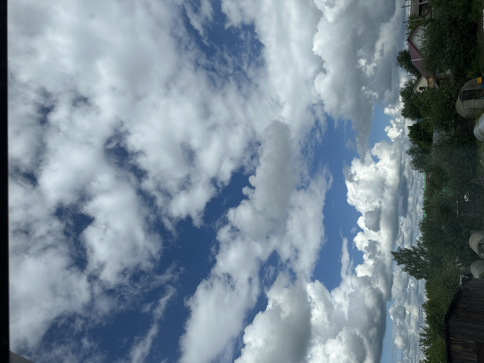 The image depicts a cloudy sky with patches of blue visible in the background. In the upper right corner, a blurred landscape is visible, including trees, a house with a red roof, and what appears to be a greenhouse or other structure. The sky is filled with various types of clouds, including cumulus clouds. The location is Unknown, Unknown.