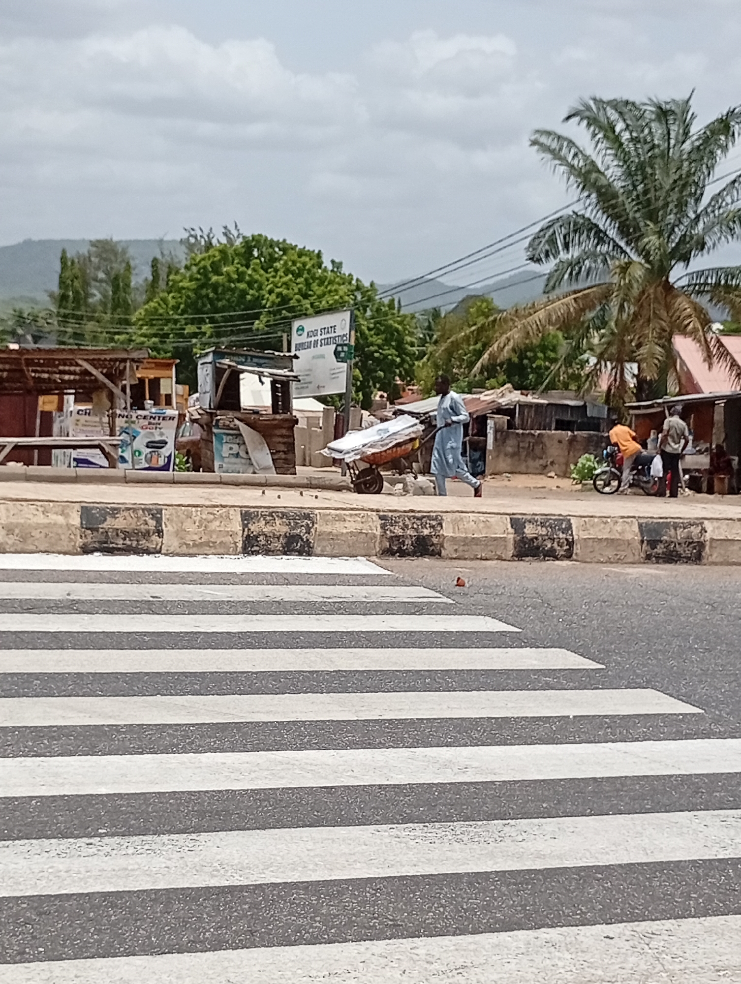 A man wearing a light blue long-sleeved shirt and dark pants is pulling a loaded handcart along a street in Lokoja, Nigeria. He is walking on the sidewalk next to a crosswalk, which is in the foreground of the image. Behind him, on the side of the road, is a motorcycle with two people standing next to it. A palm tree stands in the background. In the middle ground, there's a structure with advertising signage for "CI Trading Center," and next to it is a partially obscured sign for the "Kogi State Bureau of Statistics." Mountains are visible in the far distance. The sky is overcast.