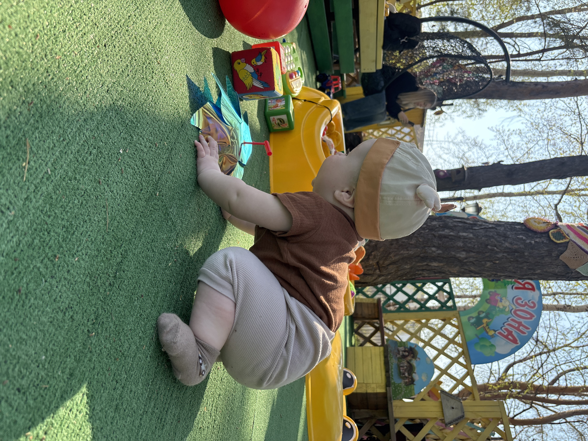 The image depicts a baby on a playground in the city of Chaikovsky, Russia. The baby is wearing a brown shirt, beige pants, and a beige hat, kneeling on green artificial turf, with a colorful pinwheel toy and building blocks nearby. Behind the baby is a yellow plastic slide and a wooden play structure decorated with cartoon images and Russian text. In the background, a woman sits in a hanging chair. The surrounding environment includes trees with budding leaves and a clear sky.