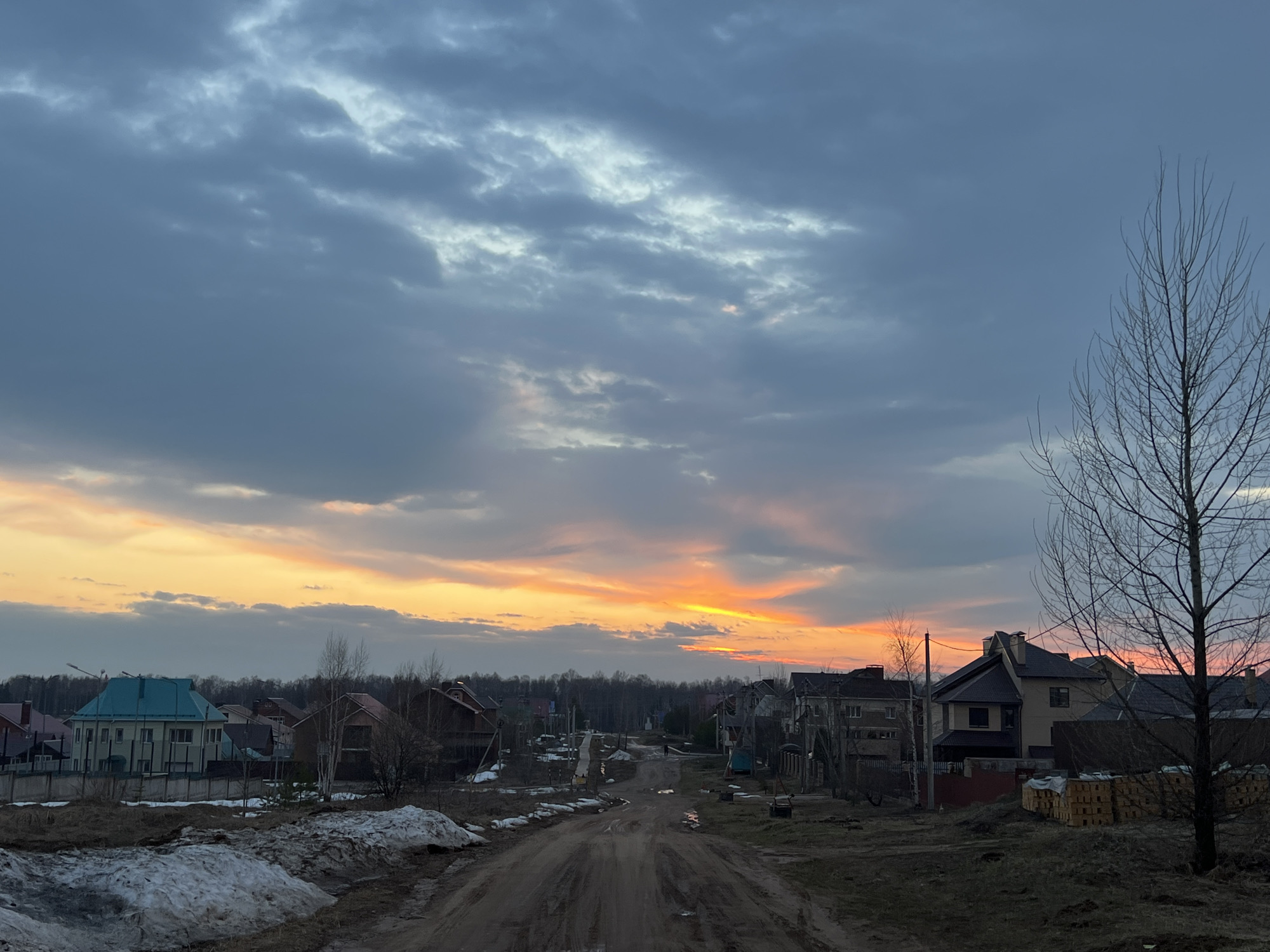 The image depicts a dirt road in Noviy, Russia, lined with residential houses, leading towards a horizon illuminated by a vibrant sunset. The sky overhead is filled with grey and white clouds. Patches of snow remain on the ground, indicating early spring or late winter. A bare tree stands to the right of the frame. Bricks are stacked near the house to the right.