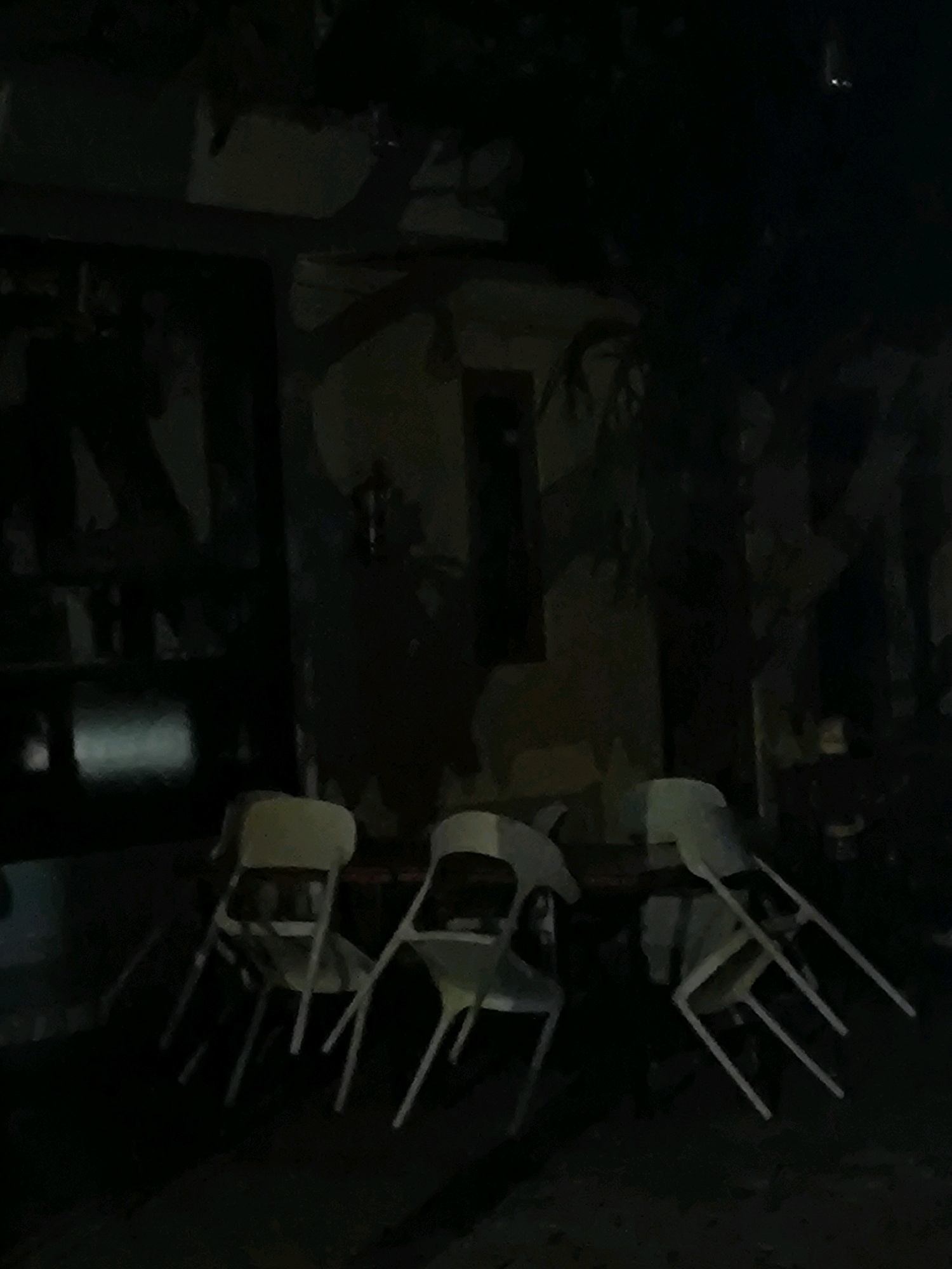 The image shows an outdoor seating area at night. There are several white chairs and a table, some of which are partially obscured by darkness. The background appears to be a wall with some sort of mural or graffiti. The overall lighting is dim, suggesting it is nighttime or a poorly lit area.