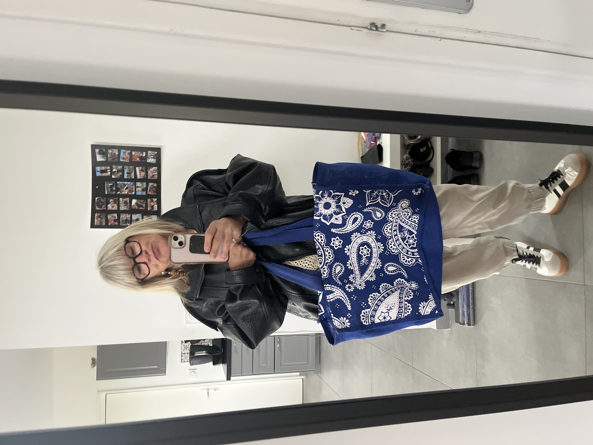 The image shows a person reflected in a mirror. The person is holding a phone in their right hand, likely taking the picture. They are wearing glasses, a black leather jacket, and tan pants. They are holding a blue bag with a paisley pattern. The reflection also shows a portion of a room with white walls, grey cabinets, a vacuum cleaner, and a collage of photos on the wall. There is also a glimpse of what appears to be a shoe rack. The city of Chambœuf, France is not discernible from the information provided.