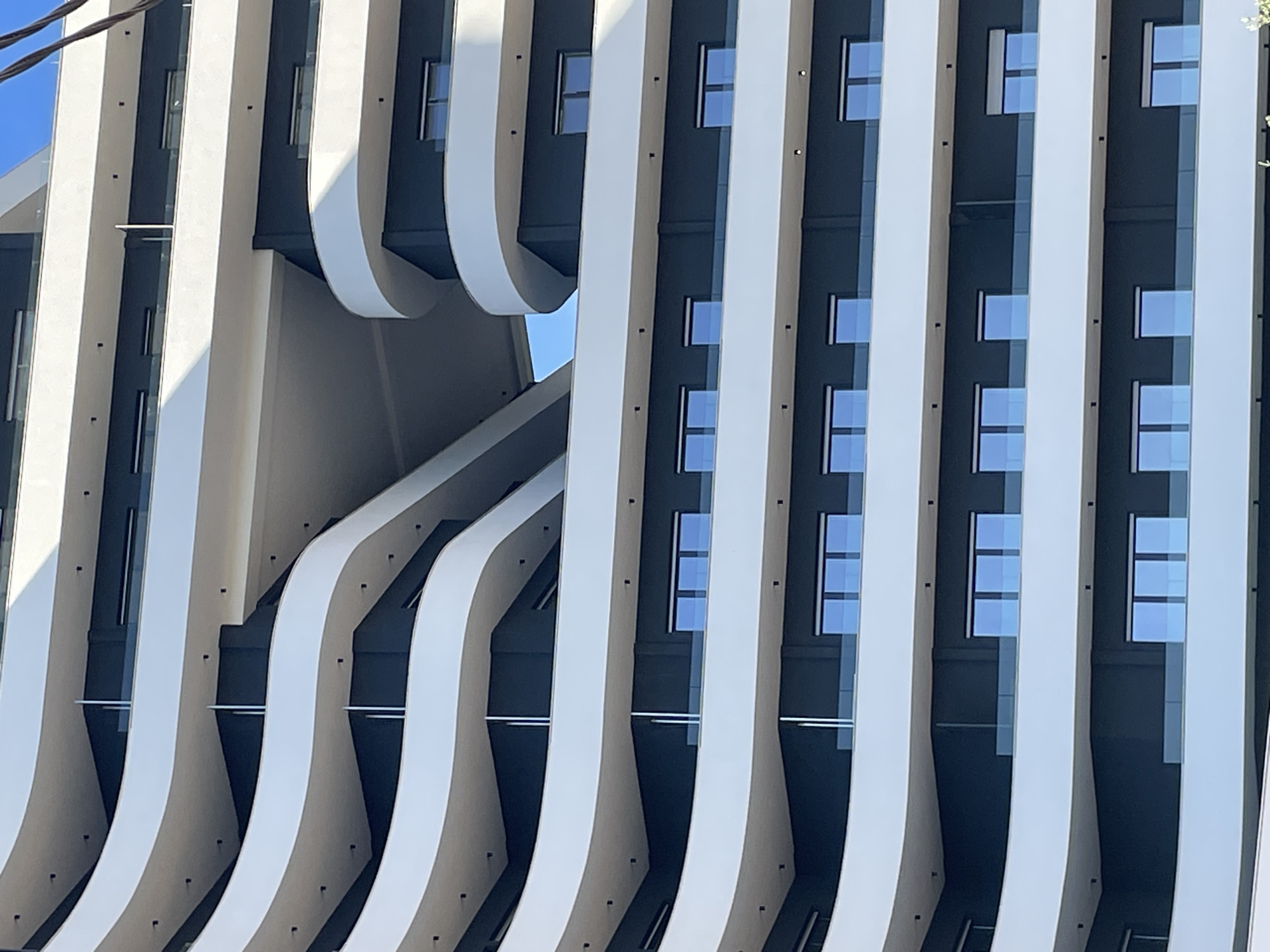 A detail shot of a modern building facade in Bar, Montenegro. The building's design features a repeating pattern of vertical white columns, some of which curve outward at the top. Dark gray or black panels are visible between the columns, and small rectangular windows are set into the panels. The sky is visible in the upper left corner. The architectural style appears contemporary and minimalist.
