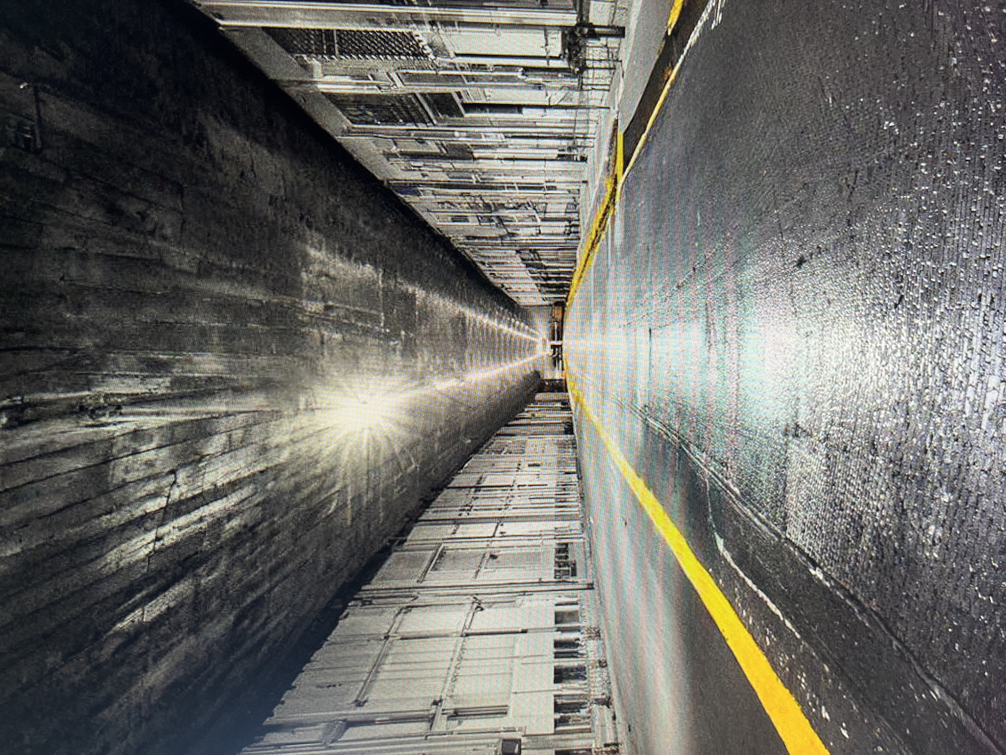 The image depicts an urban underpass or overpass with a concrete ceiling and parallel walls. A yellow line marks the center of the asphalt road, which appears wet, reflecting the light source at the end of the tunnel. The walls are lined with numerous windows or openings, suggesting a building structure adjacent to the road. The overall scene is characterized by strong perspective lines and a dramatic interplay of light and shadow, creating a sense of depth and enclosure. No people or vehicles are visible.