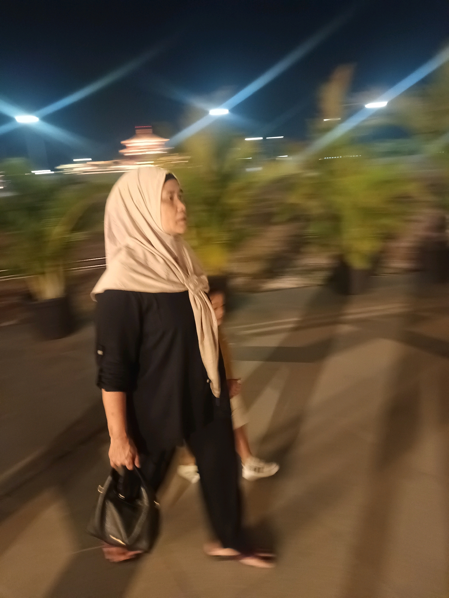 A woman wearing a light brown hijab and a black abaya is walking outdoors at night. She is holding a black handbag in her right hand. Behind her, a blurred figure of a child wearing white shoes is visible. The background shows blurred green foliage and streetlights. The image appears to be taken in North Jakarta, Indonesia, based on the characteristic building visible in the far background.