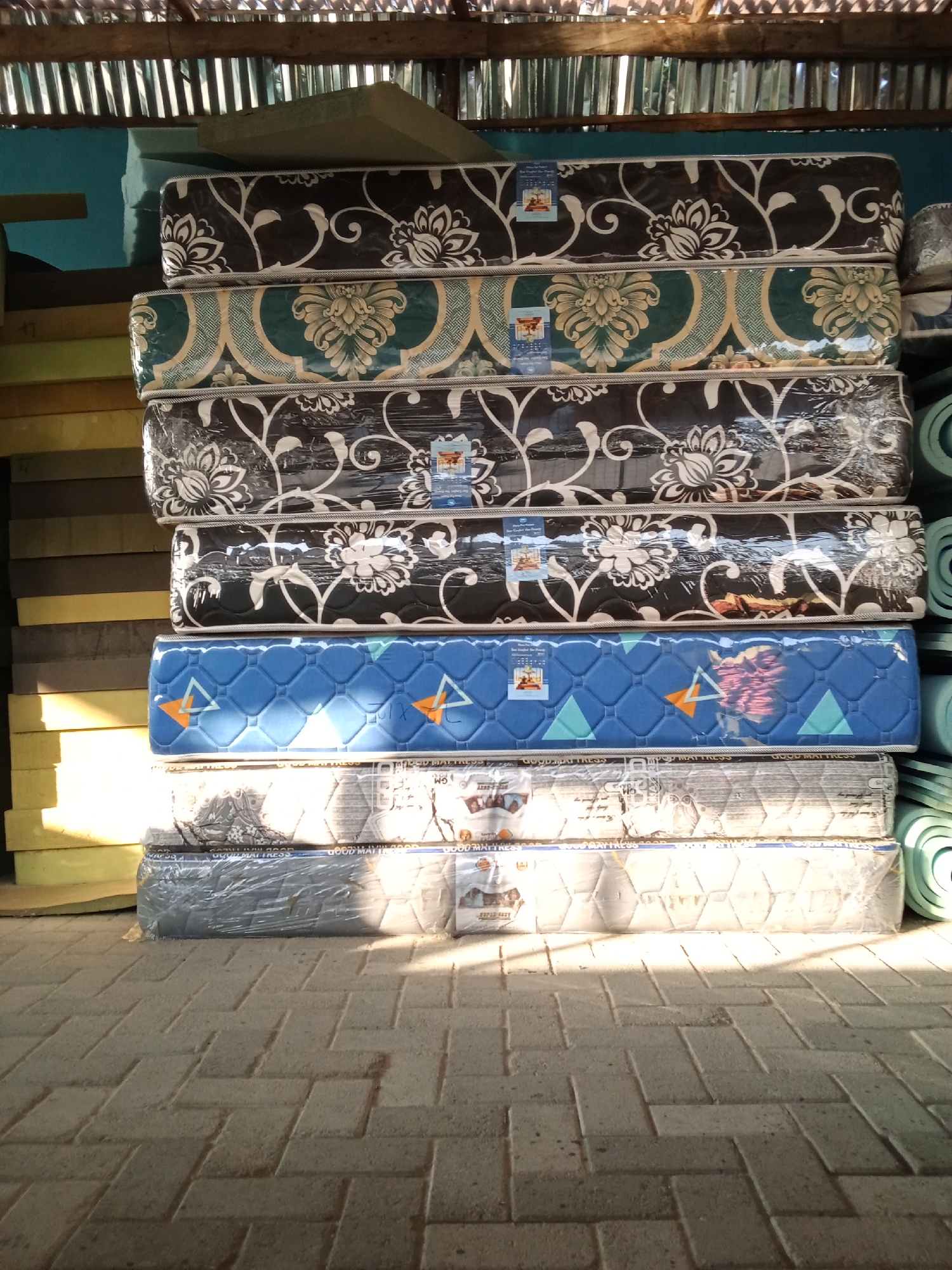 The image shows a stack of mattresses in what appears to be a storage area or shop in Ado, Nigeria. The mattresses are wrapped in clear plastic and have various patterns. The top mattress has a black background with white floral designs. Below it is a mattress with a green and gold pattern. The next mattress down also has a black background with white floral designs. The fourth mattress from the top is blue with a diamond pattern and orange triangles. The fifth mattress has a patterned fabric with white and gray tones. The bottom mattress is wrapped in gray plastic. In the background, there are other stacked materials, possibly foam or more mattresses, and a corrugated metal roof overhead. The floor is paved with interlocking bricks, and shadows from the sunlight are visible on the floor. The scene suggests a commercial setting where mattresses are stored or sold. There are no people visible in the image.