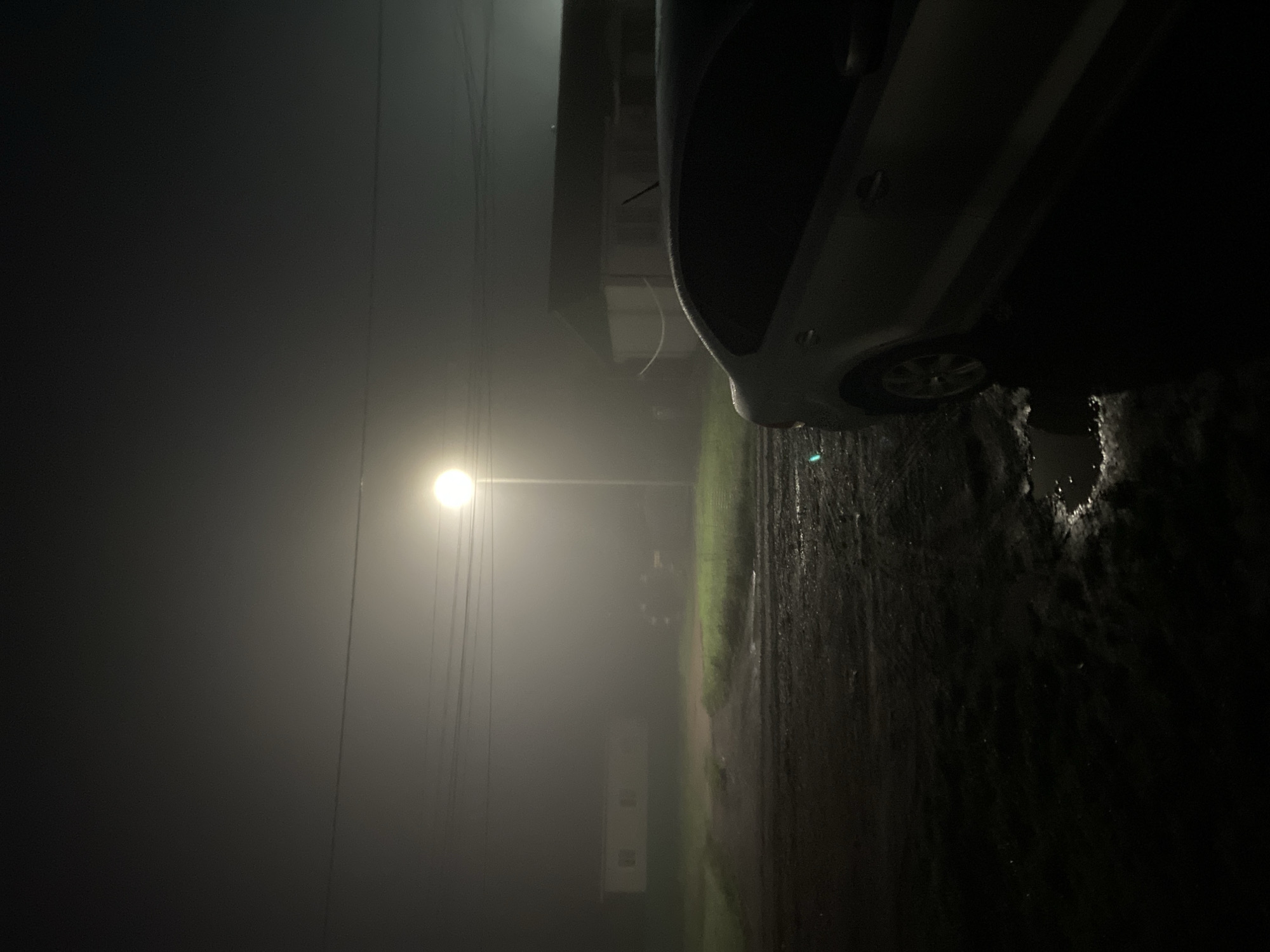 In Chirsha-Tartysh, Russia, a silver sedan is parked on a dark, wet, and muddy road at night. The scene is obscured by a thick fog that casts a dim, diffused light, illuminated by a solitary street lamp positioned in the mid-ground. The lamp's glow is softened by the fog, creating a hazy atmosphere. Behind the parked car and to the left, a low-lying building with visible windows is partially visible through the mist. To the right of the street lamp, another building is dimly outlined against the foggy sky. Power lines are visible crisscrossing the upper portion of the frame, disappearing into the fog. The ground in front of the car appears to have puddles of water, reflecting the ambient light.