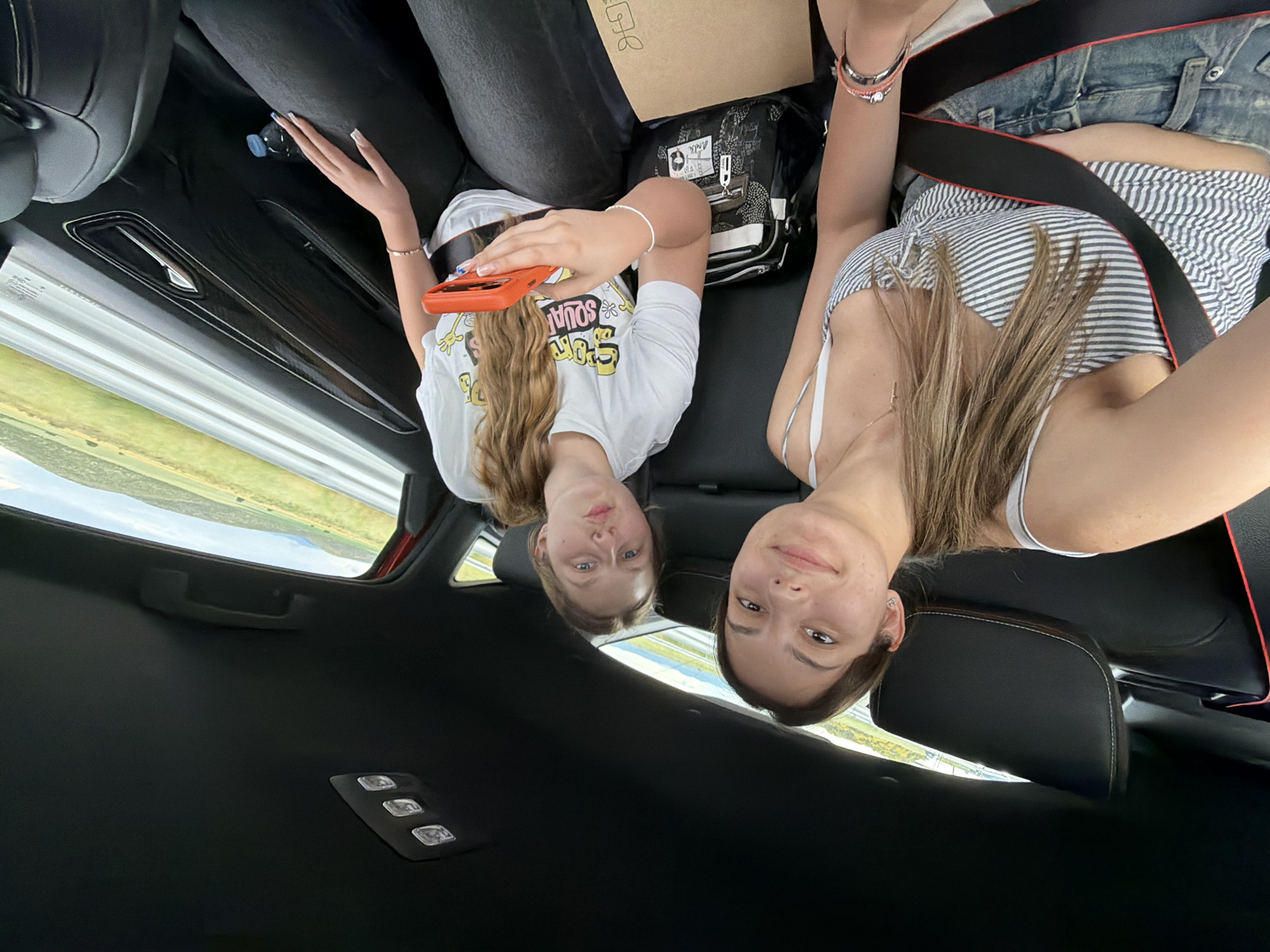 Two young women are in a vehicle. One woman is wearing a white t-shirt with a graphic, holding an orange cell phone. The other woman is wearing a striped tank top and denim shorts. Both women are wearing seatbelts. The environment outside the vehicle shows a landscape with greenery, viewed through the windows. There is no indication to verify if the location is Koniovo, Bulgaria.