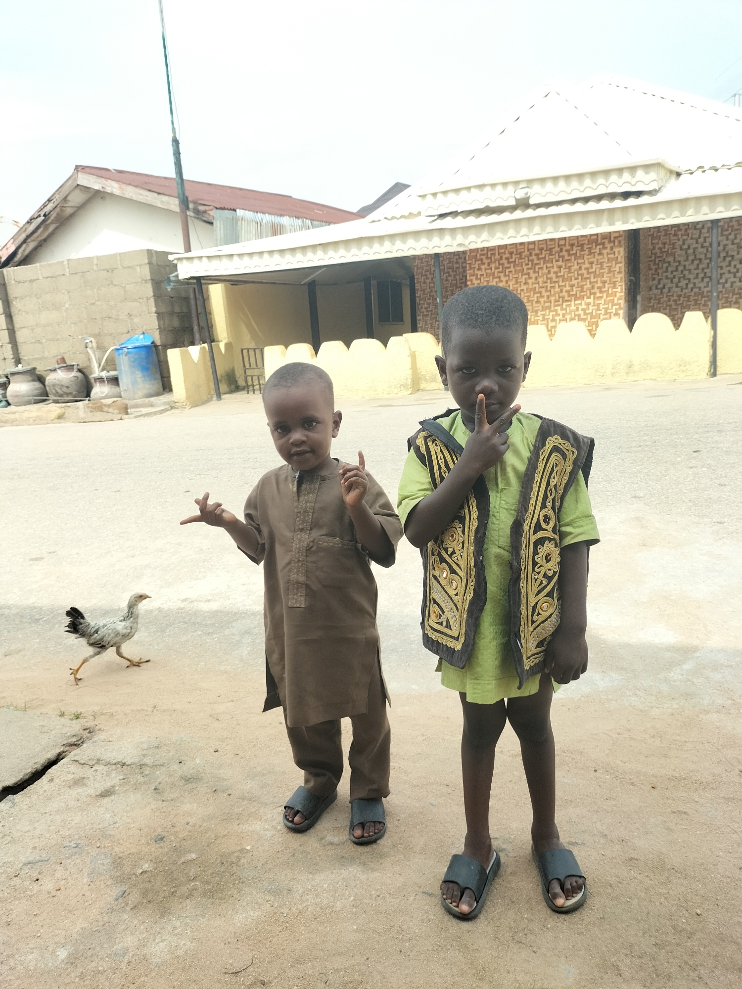 Two young boys stand on a dusty, cracked road in Jalingo, Nigeria. The boy on the left, wearing a brown robe, gestures with his left hand. To his left, a young chicken walks away from the camera. The boy on the right, dressed in a green outfit and an ornate black and gold vest, holds up two fingers in a peace sign. Behind them, a low, light-yellow wall with decorative indentations separates the road from a building with a white roof and a textured brown facade. In the background, a tall pole stands next to buildings with corrugated metal and tiled roofs. A blue plastic water barrel and several large clay pots are visible near a brick wall on the left side of the image.