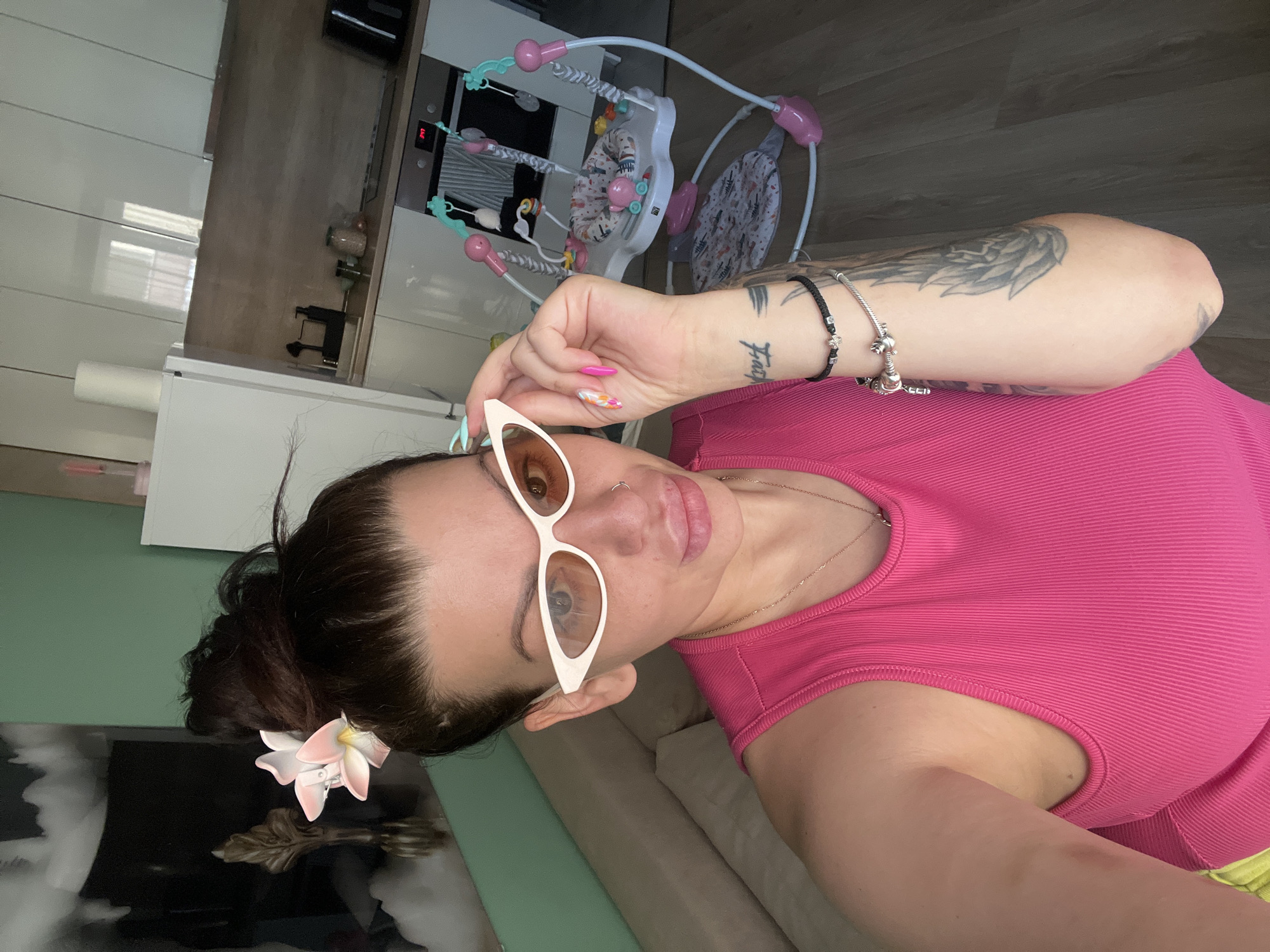 A woman with dark hair pulled up in a messy bun, adorned with a pink and white flower, is taking a selfie. She is wearing white cat-eye sunglasses with orange lenses, a bright pink ribbed tank top, and a delicate gold necklace. Her lips are full, and she has a nose ring. On her right arm, she has several tattoos and is wearing two bracelets, one black with a silver charm and another silver charm bracelet. Her fingernails are painted with a colorful, abstract design. She is holding the edge of her sunglasses with her right hand, with her thumb and index finger visible.
The background suggests an indoor domestic setting, likely an apartment. To her left, a cream-colored sofa is visible. Behind her, a kitchen area is partially visible, featuring white cabinets, a light brown countertop, a sink with a black dispenser, and a stovetop with a digital display showing "3:47". A white refrigerator stands to the far left of the kitchen area. In the middle ground, a baby jumper or activity center with pink and blue accents is set up. The flooring is a dark wood laminate. The overall lighting indicates it is daytime, with sunlight entering the room. The location is described as Plovdiv, Bulgaria.