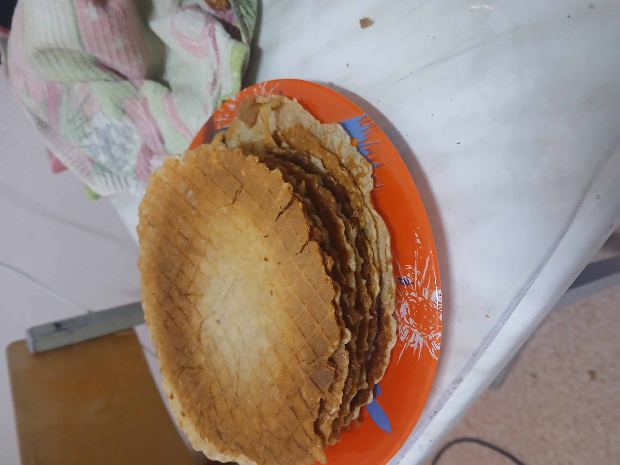 The scene shows a stack of waffle cookies on an orange plate with a white floral and blue flame-like design around the rim. The plate rests on a white, potentially plastic, table or surface. To the left of the plate, a dishtowel with pink, green, and white floral patterns is visible. A small section of a wooden cutting board is also present at the bottom left corner of the frame. The location is in a residential or domestic setting, likely in the city of Chaikovsky, Russia, based on the user's request.
